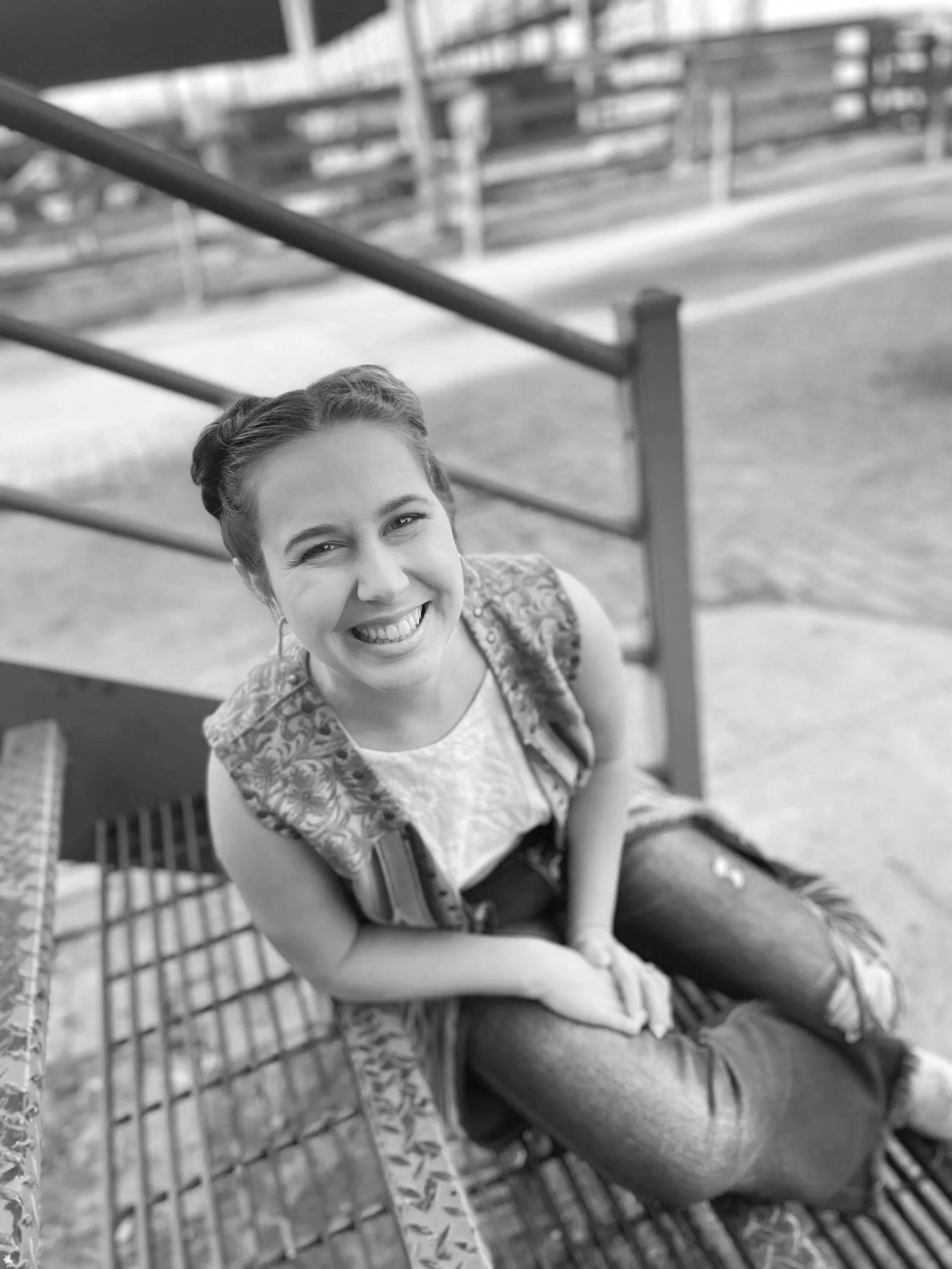 A young woman with a wide smile, sitting on a metal bench outdoors at a park, looking up at the camera.
