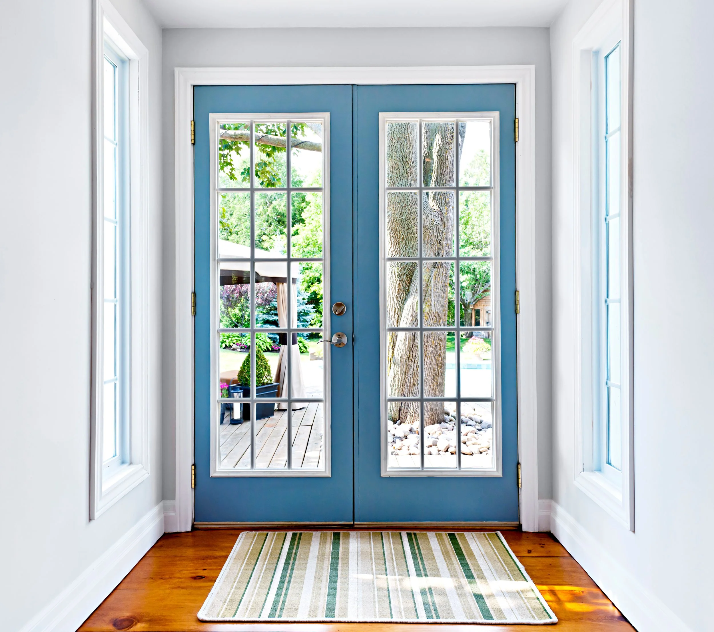 View of a front door with glass panes leading outside to a garden with trees and patio, flanked by two side windows, with a striped rug on a wooden floor inside.