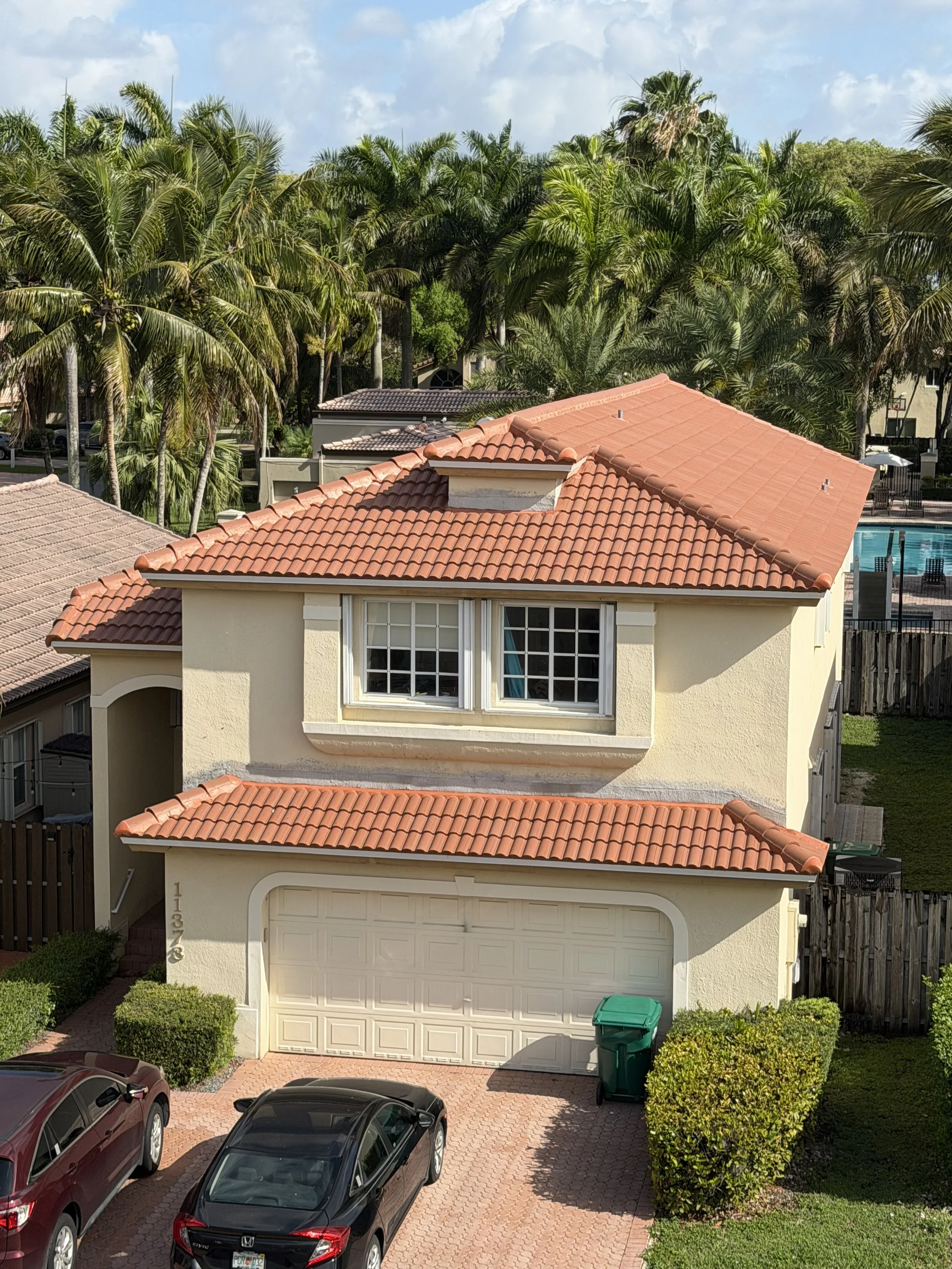 A two-story house with a red tiled roof, cream-colored exterior walls, and a white garage door. There are two cars parked in the driveway and a green trash bin near the bushes. Tall palm trees and other greenery surround the house, with a swimming pool visible in the background.