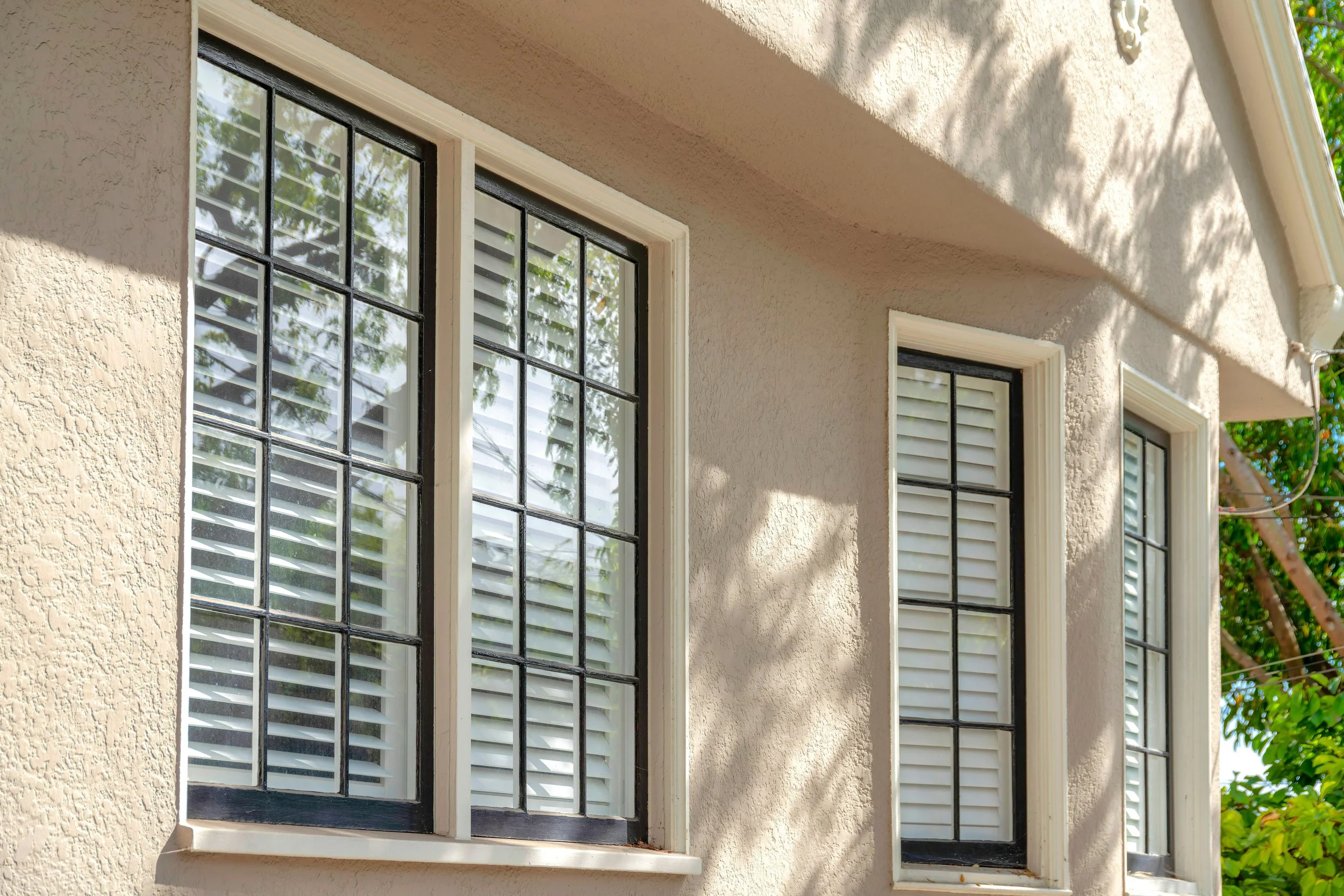 Three windows with black grid frames and white shutters on a beige stucco exterior wall of a house, with shadows of tree branches on the wall.