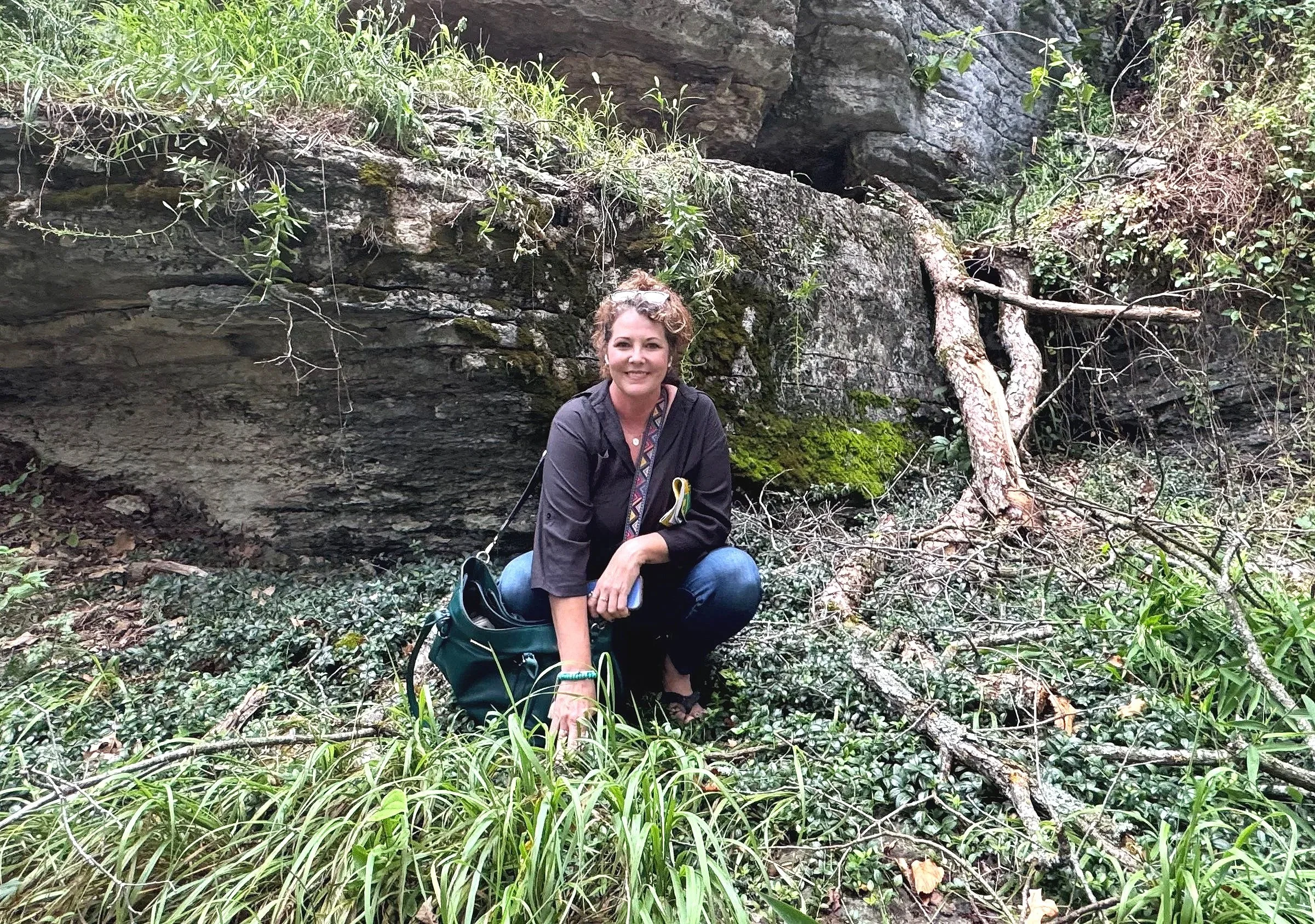 A woman with curly hair squatting outdoors in a natural green area with rocks, plants, and fallen branches.