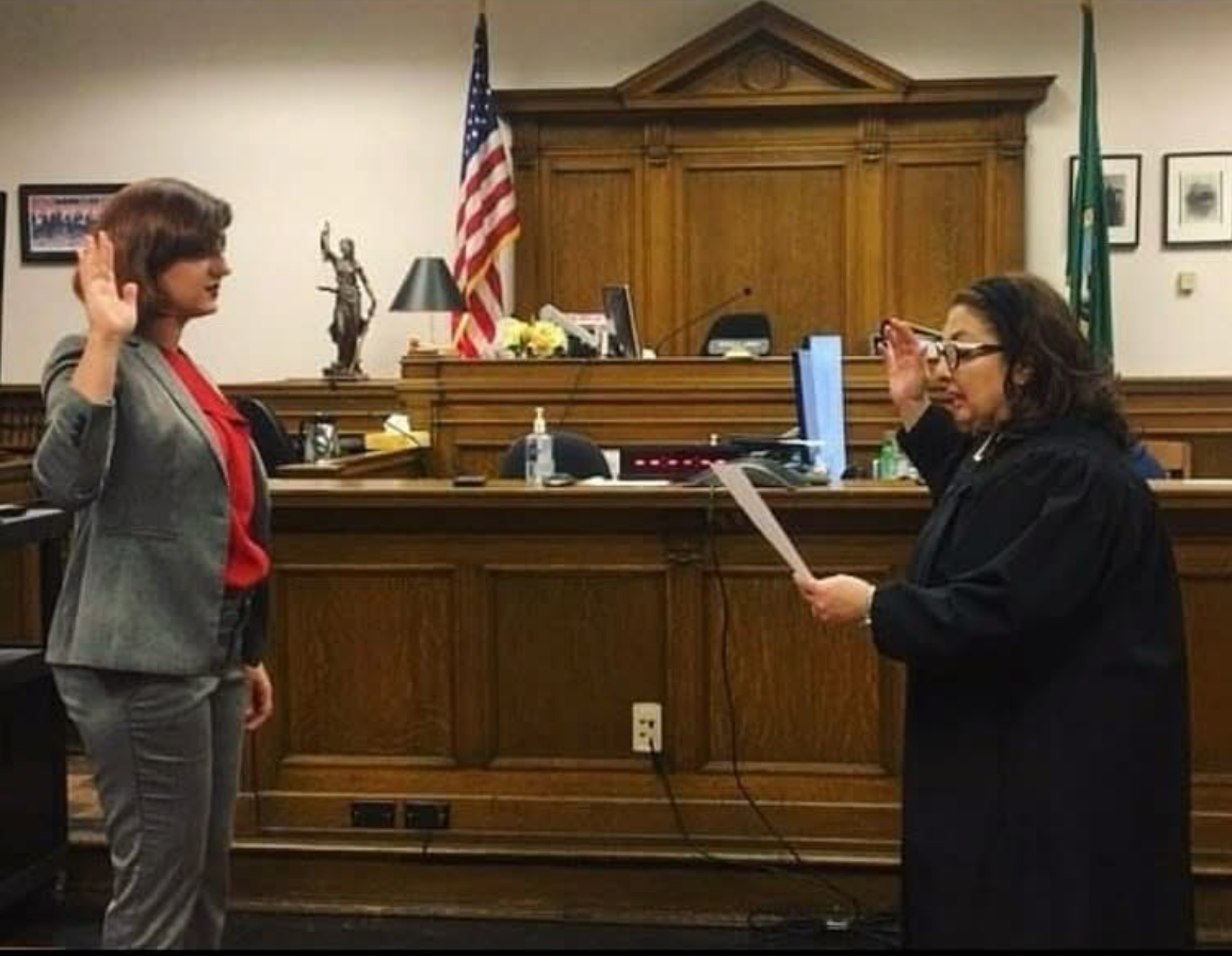 A woman taking the oath of office in a courtroom with her hand raised, while another woman in a black robe reading from paper.