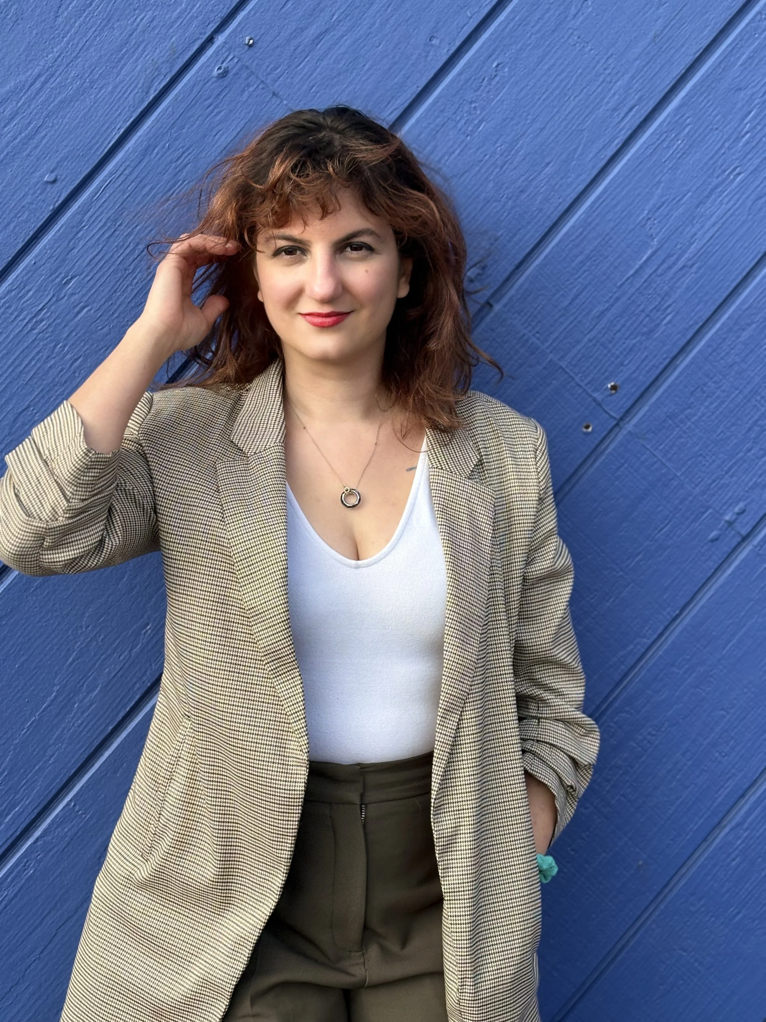 A woman with curly reddish-brown hair, wearing a plaid blazer over a white top, standing against a blue wooden wall. She has a necklace and is smiling at the camera.
