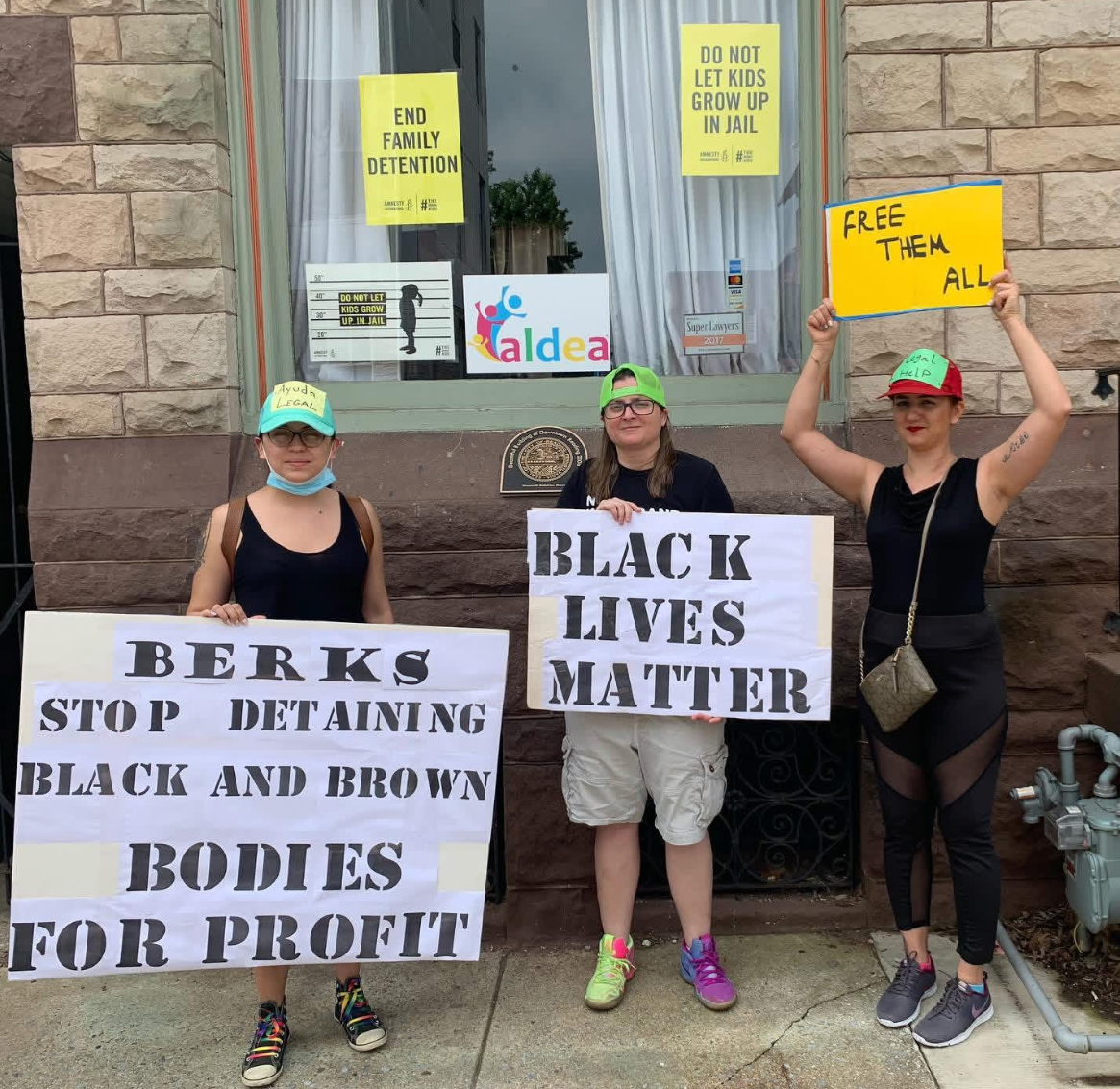 Three women protesting outside a building, holding signs advocating for Black lives and criticizing family detention. The woman on the left wears a blue cap with a yellow sign reading 'Ayuda Legal' and a face mask, holding a sign that says 'Berks stop detaining black and brown bodies for profit.' The woman in the middle wears a green cap and glasses, holding a sign that says 'Black lives matter.' The woman on the right wears a red cap with a green sign reading 'Legal Help,' holding a sign that says 'Free them all.' The window behind them has signs including 'End family detention' and 'Do not let kids grow up in jail,' and a colorful logo that reads 'aldEa.'