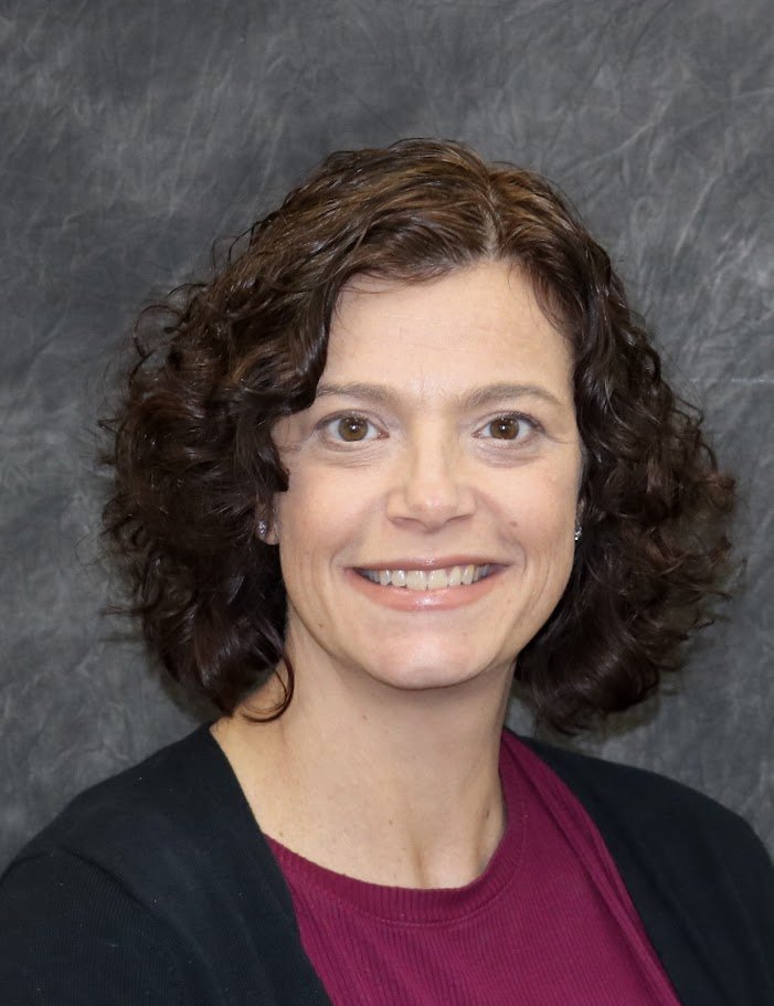 A woman with short curly brown hair, smiling, wearing a black top over a maroon shirt, against a dark marble background.