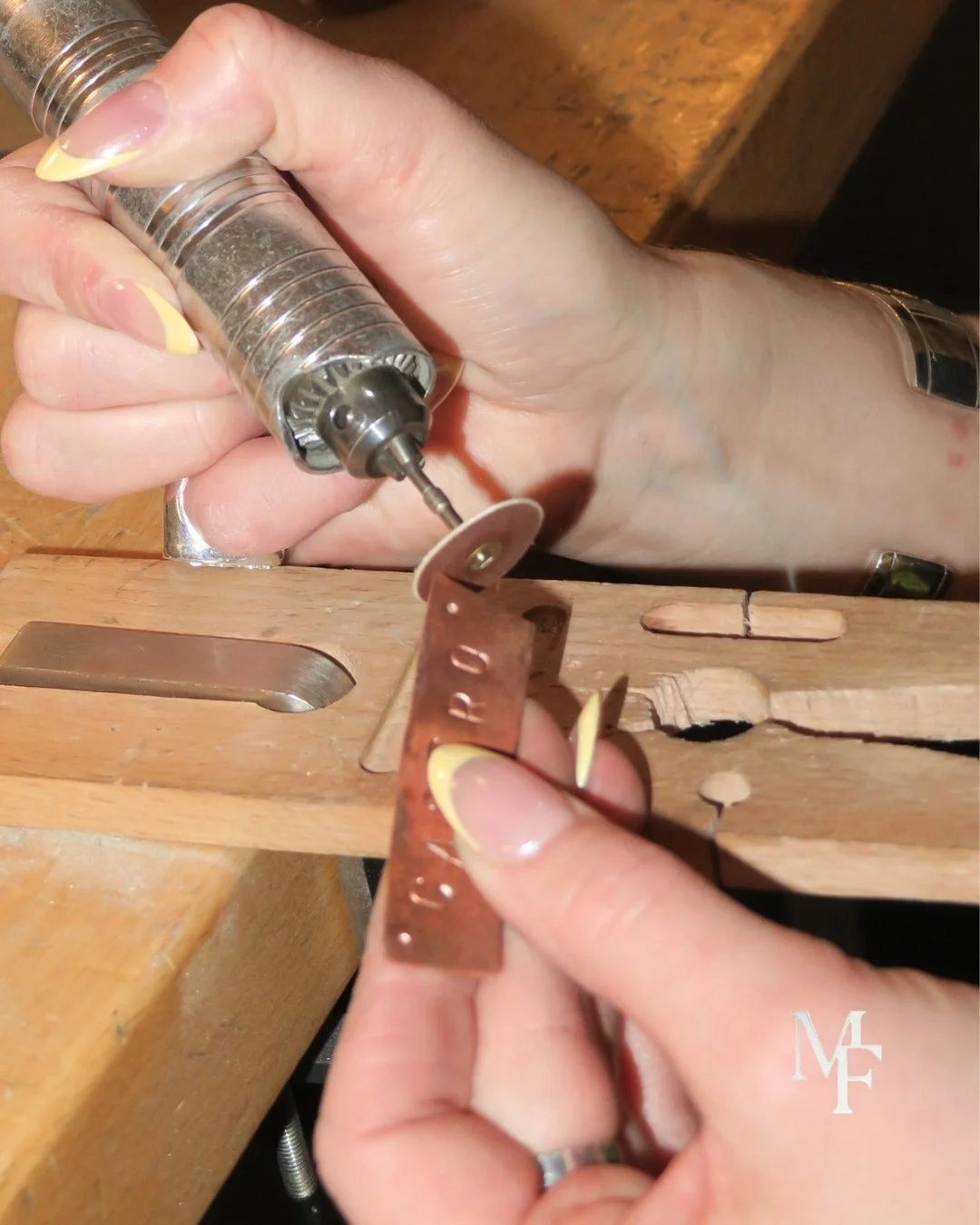 Close-up of a person using a rotary tool with a disc attachment to carve a small piece of copper or brass metal. The work is being done on a wooden workbench.