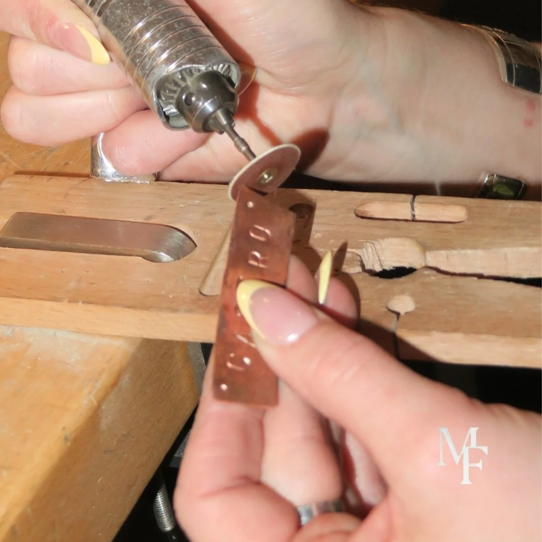 Close-up of hands holding a small rotary tool and a metal measuring strip, using the rotary tool to cut or shape the metal strip on a wooden workbench.