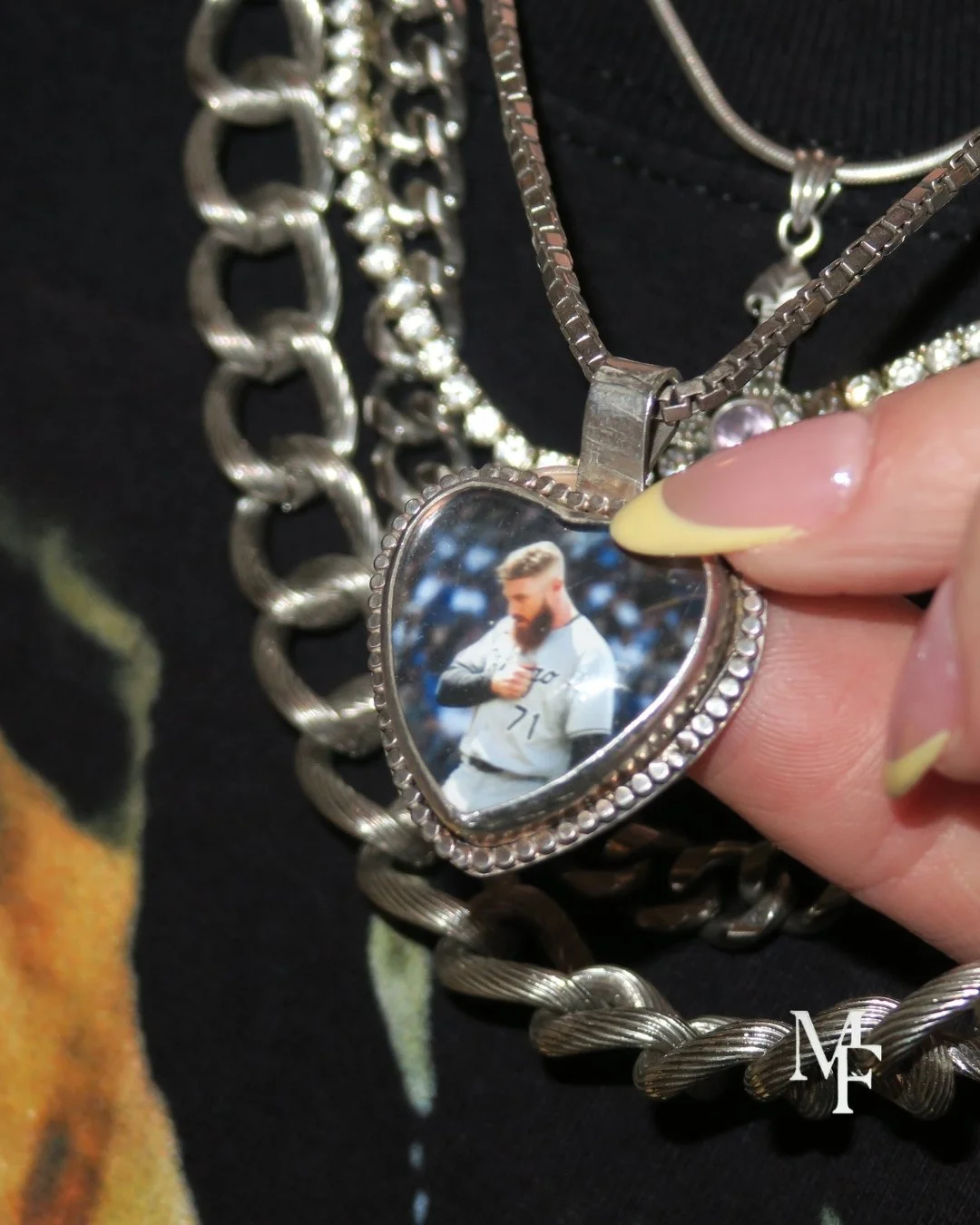 A close-up of a silver necklace with a heart-shaped pendant containing a photo of a bearded man in a white sports uniform. The person holding the pendant has long, yellow-tipped nails and a hand with neatly manicure. Multiple chunky silver chains are layered around the pendant.