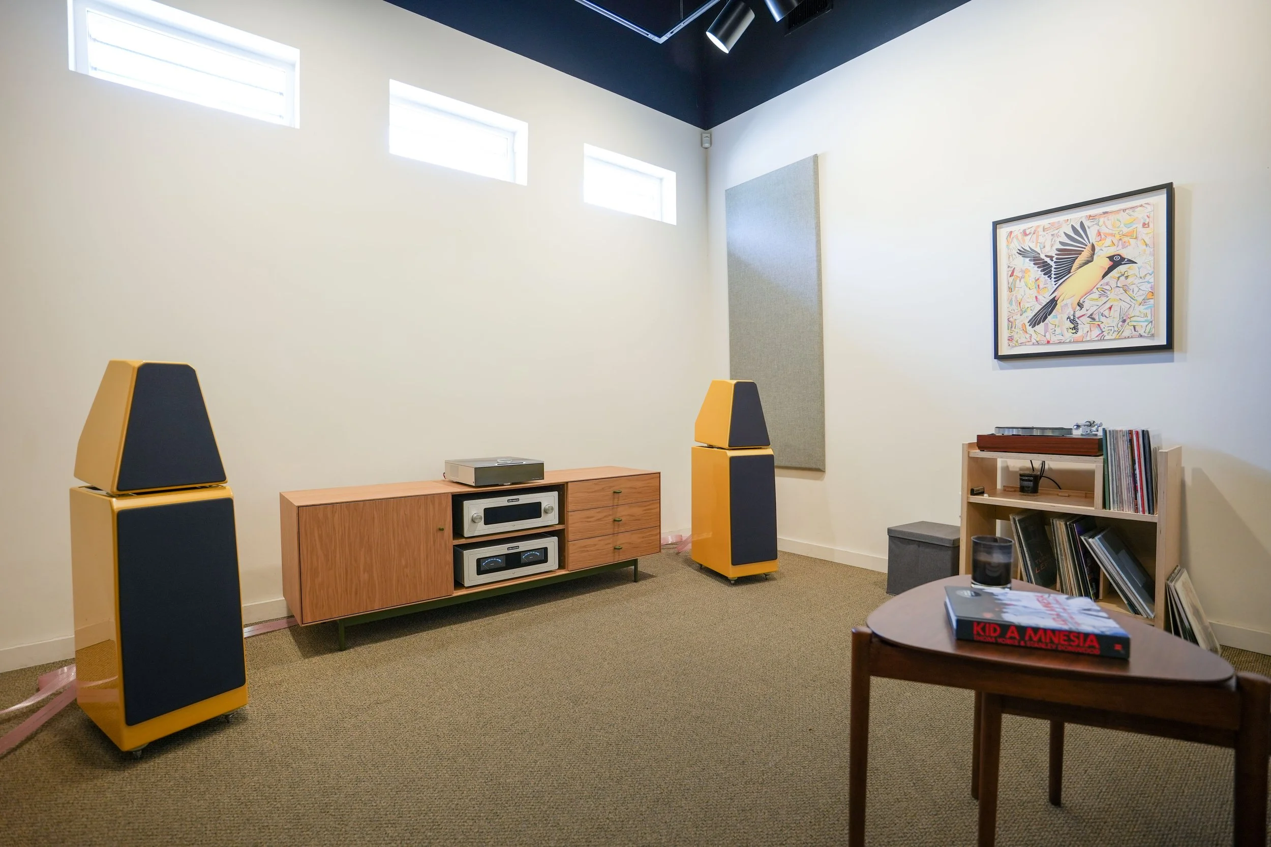 A minimalistic living room with high small windows, a framed bird illustration on the wall, vintage audio equipment on a wooden sideboard, and yellow and black speakers. A small wooden table with a book, glass, and remote control is also visible.