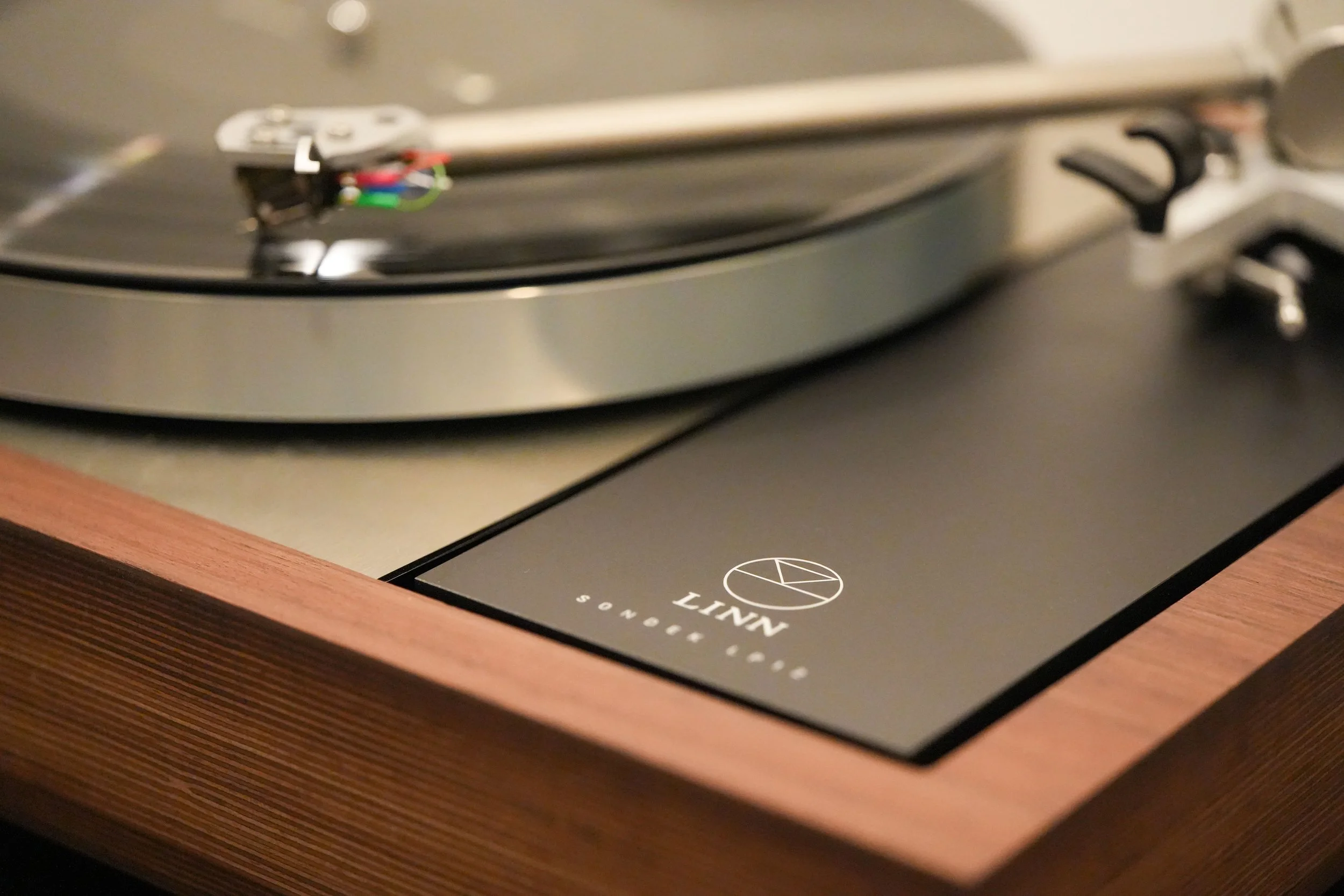 Close-up of a turntable with a wooden base and a black platter, showing the tonearm and stylus in a resting position.