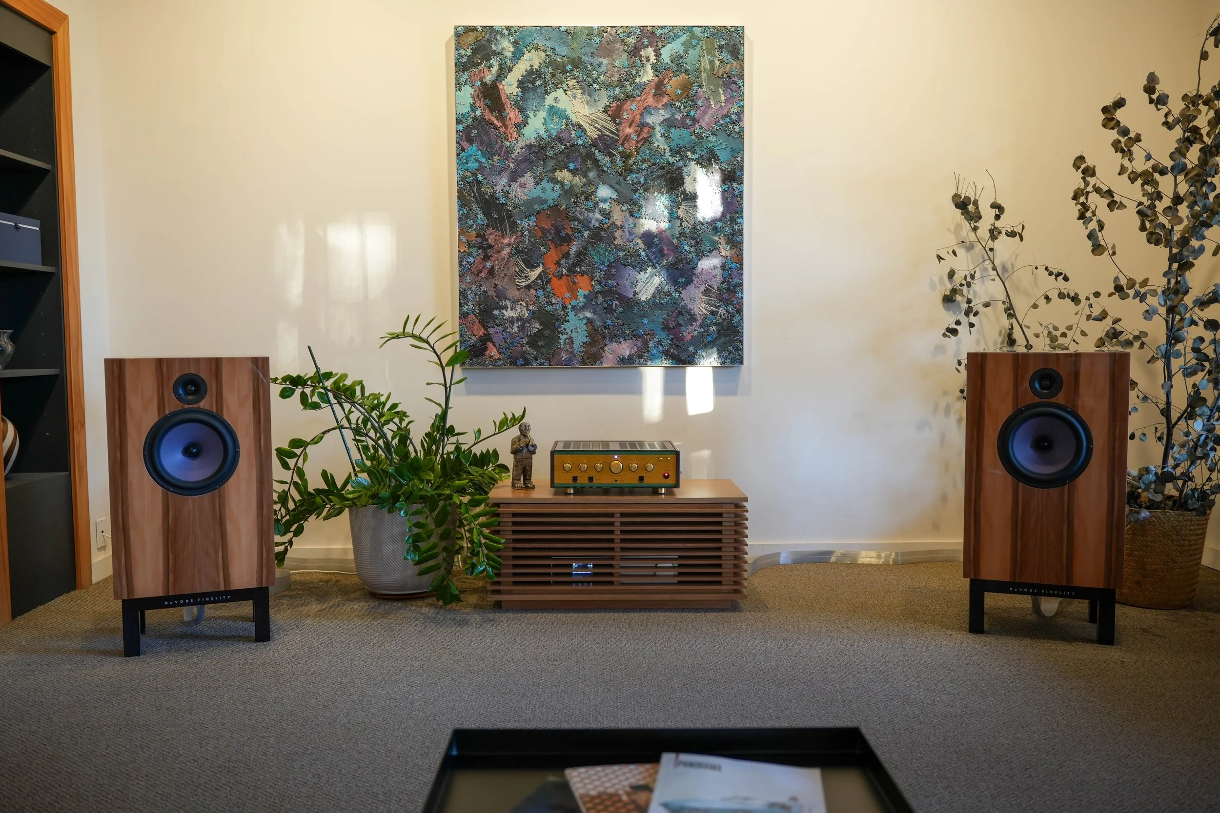 A living room with two large wooden speakers on black stands, a potted plant on the left, a vintage audio amplifier on a wooden console in the center, a colorful abstract painting above, and tall leafy plants on the right.