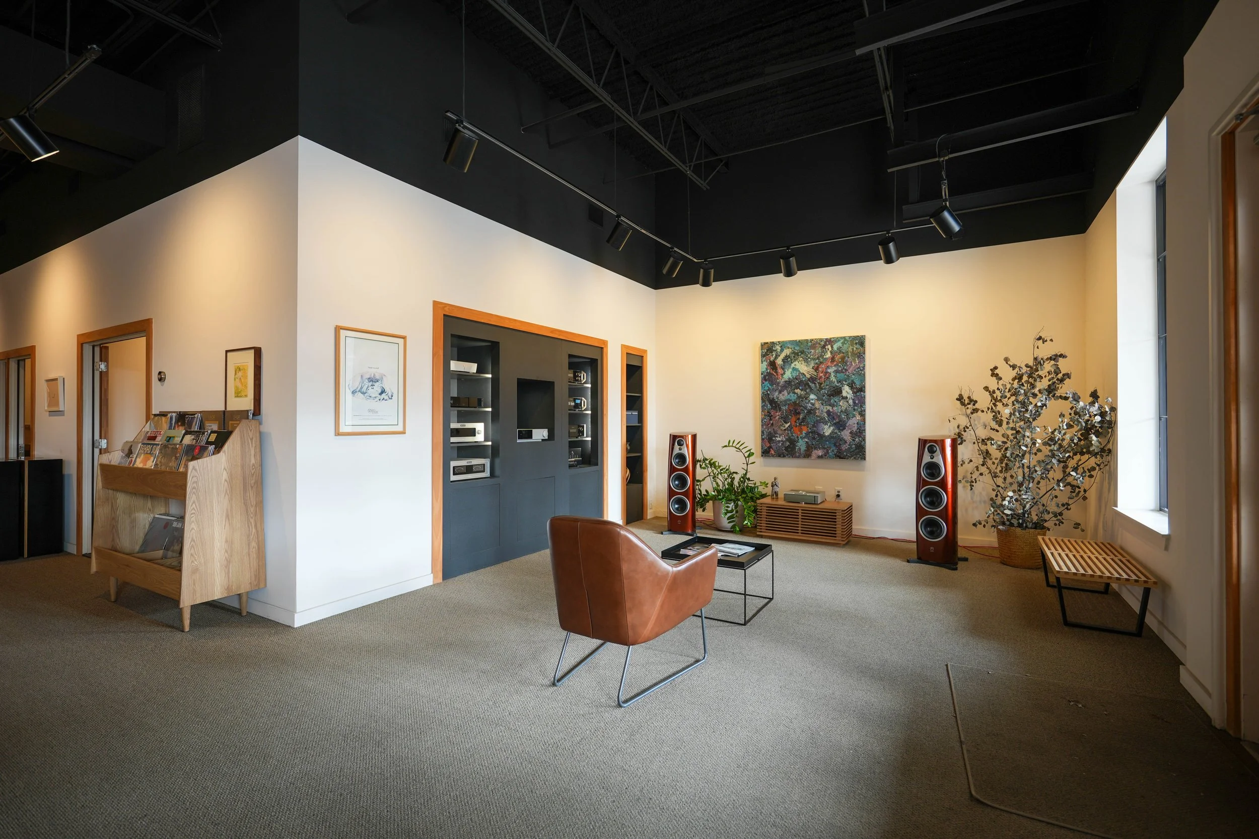 Interior of a modern living room or lounge with a brown leather chair, black coffee table, two large speakers, artwork, plants, and a window.