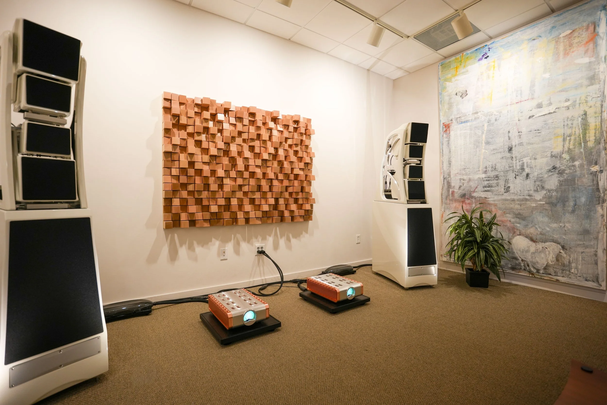 A room with acoustic panels and high-end audio equipment, including large speakers, amplifiers, a colorful abstract painting, and a potted plant.