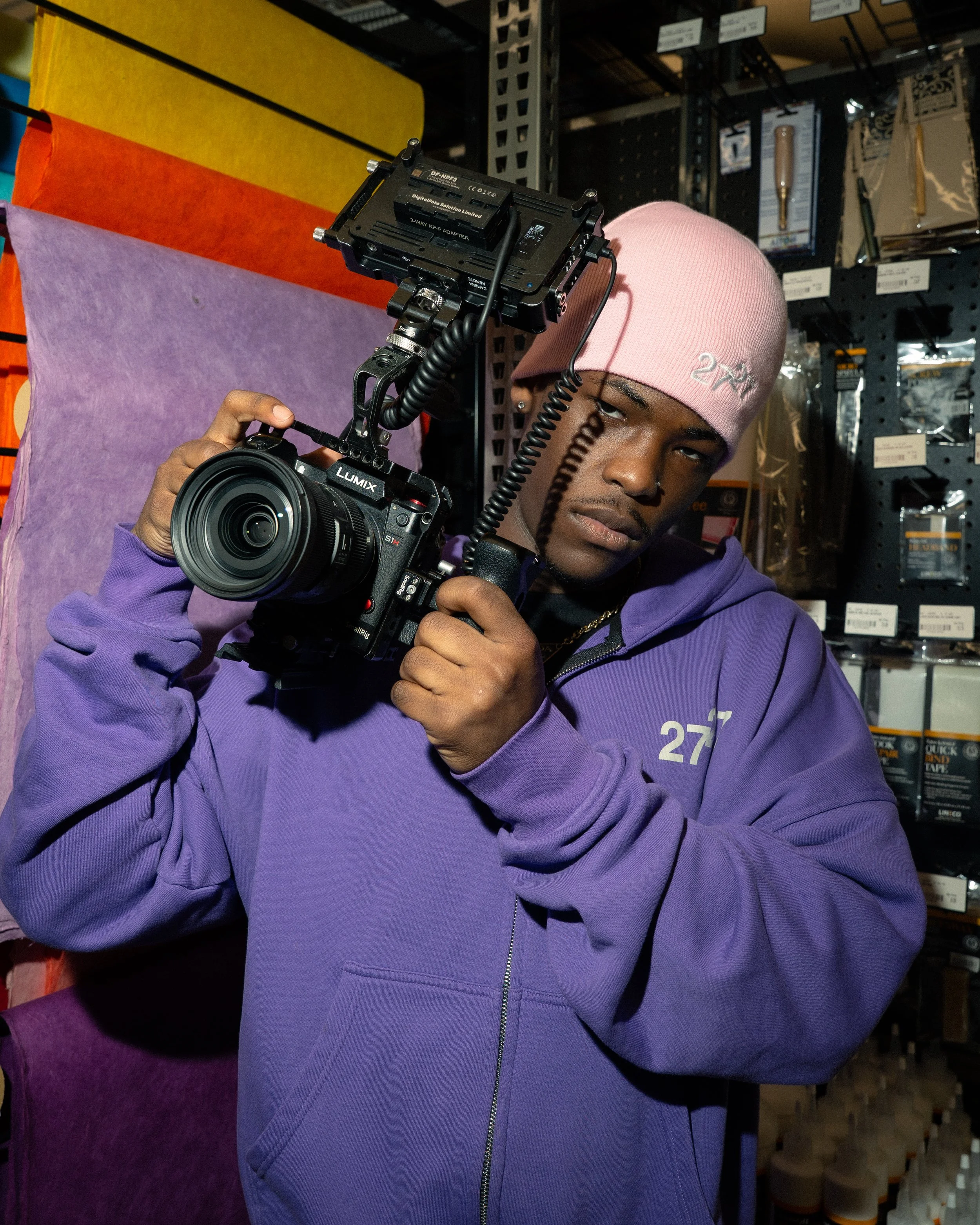 A young man in a pink beanie and purple hoodie holding a professional camera is in a store aisle surrounded by colorful fabrics and home repair supplies.