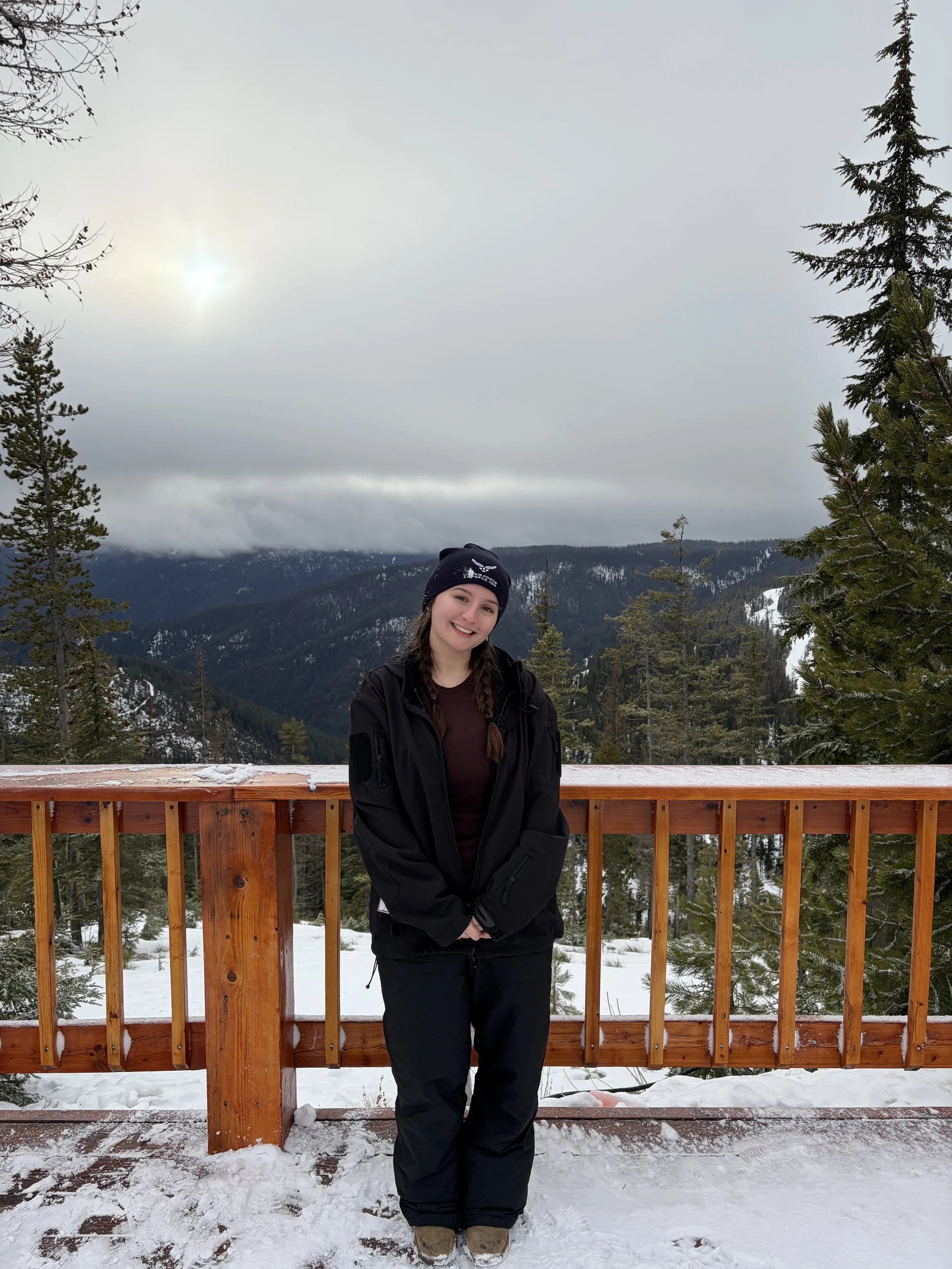 A woman dressed in winter clothing standing on a snow-covered deck with a mountain landscape in the background, cloudy sky, and evergreen trees.