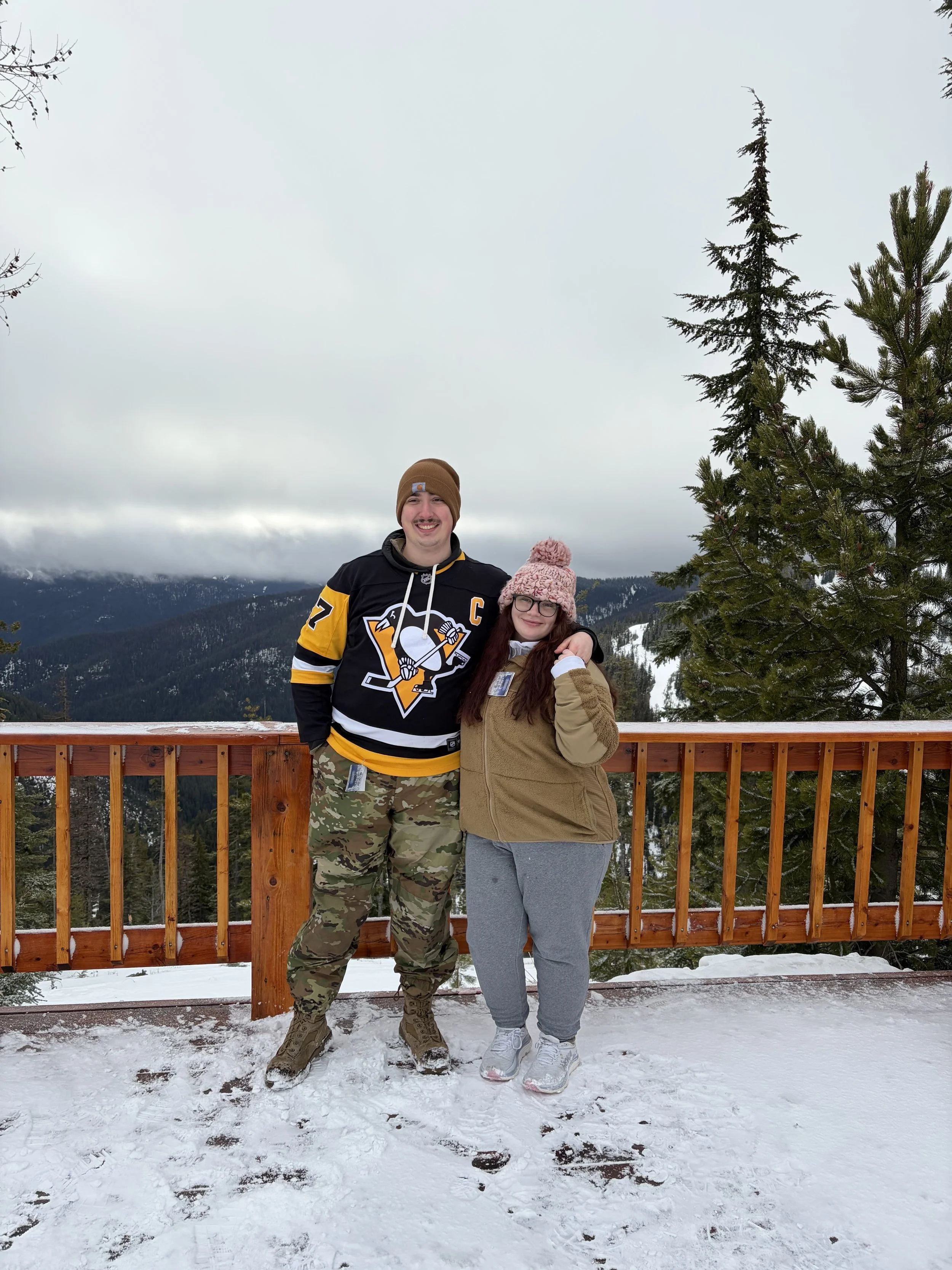 A man and a woman standing together on a snowy wooden deck outdoors, surrounded by pine trees and mountain scenery, with the man wearing a Pittsburgh Penguins hockey jersey and camouflage pants, and the woman wearing a tan fleece jacket and gray sweatpants, both smiling and embracing.