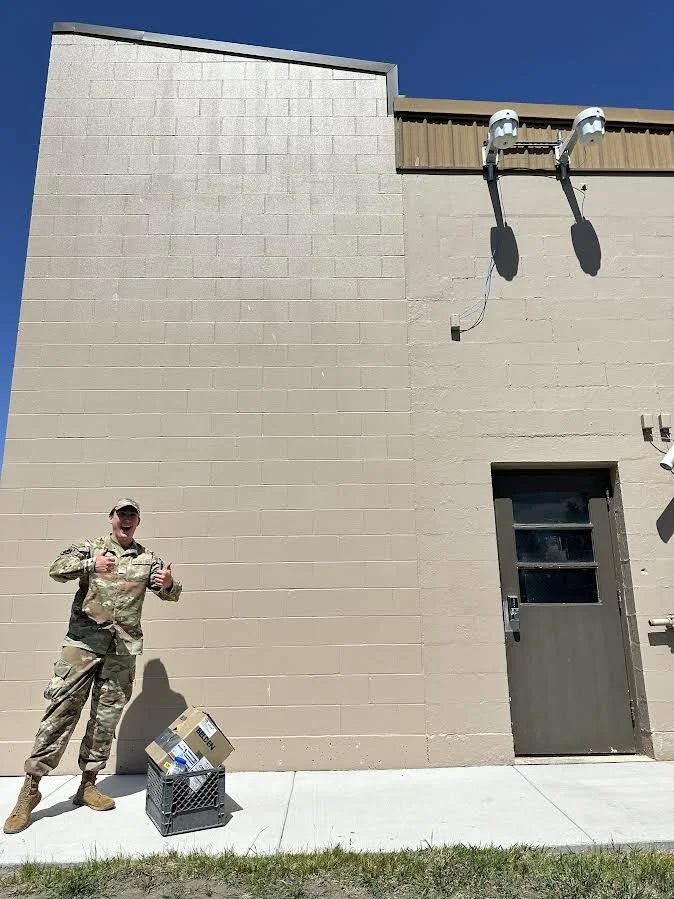 A man in military camouflage uniform standing outside a beige building giving a thumbs up. There is a shopping basket with boxes on the ground next to him. The building has a door and two mounted security cameras with shadows cast on the wall.