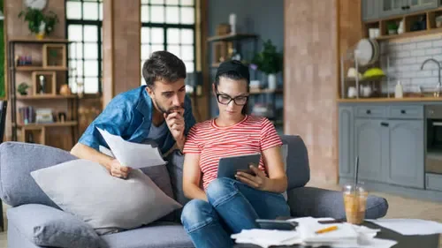 A man and woman sitting on a gray sofa in a modern living room, looking at a tablet and holding papers, with a coffee cup on the table.