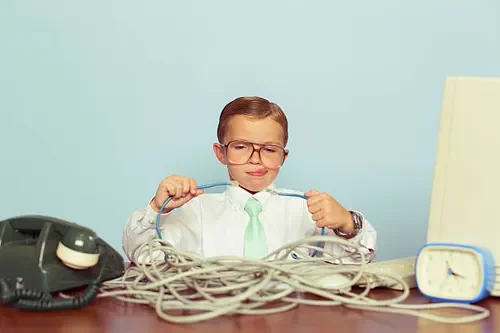 Child boy with glasses sitting at desk tangled in telephone cords, looking frustrated, with a landline phone, clock, and whiteboard in background.