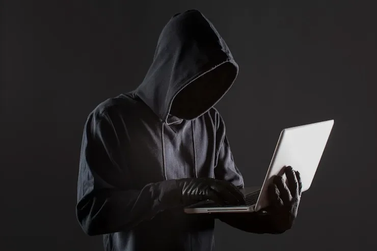 A person in a dark hoodie with the face hidden, typing on a laptop against a dark background.
