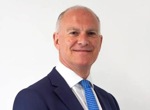 Smiling middle-aged man in a navy suit, white shirt, and blue tie, posing against a plain white background.