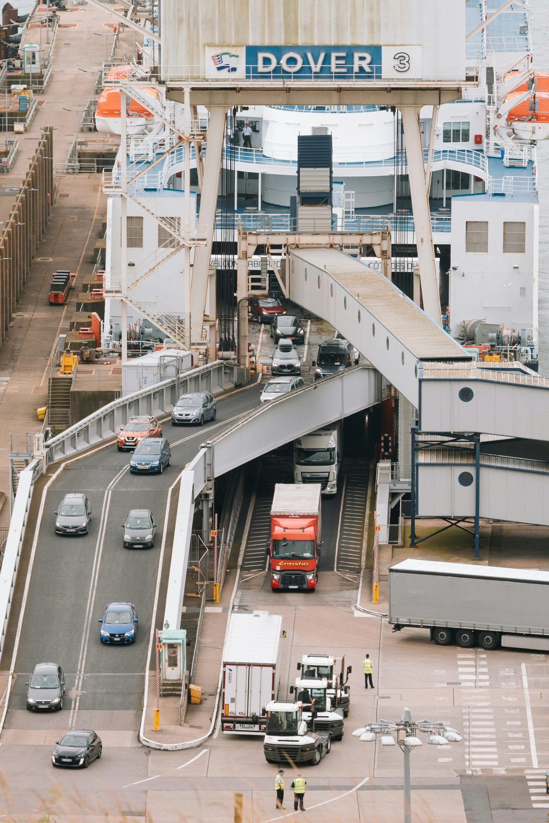 A large ferry boat docked at a port, with cars driving on and off through a ramp, and workers in safety vests nearby.