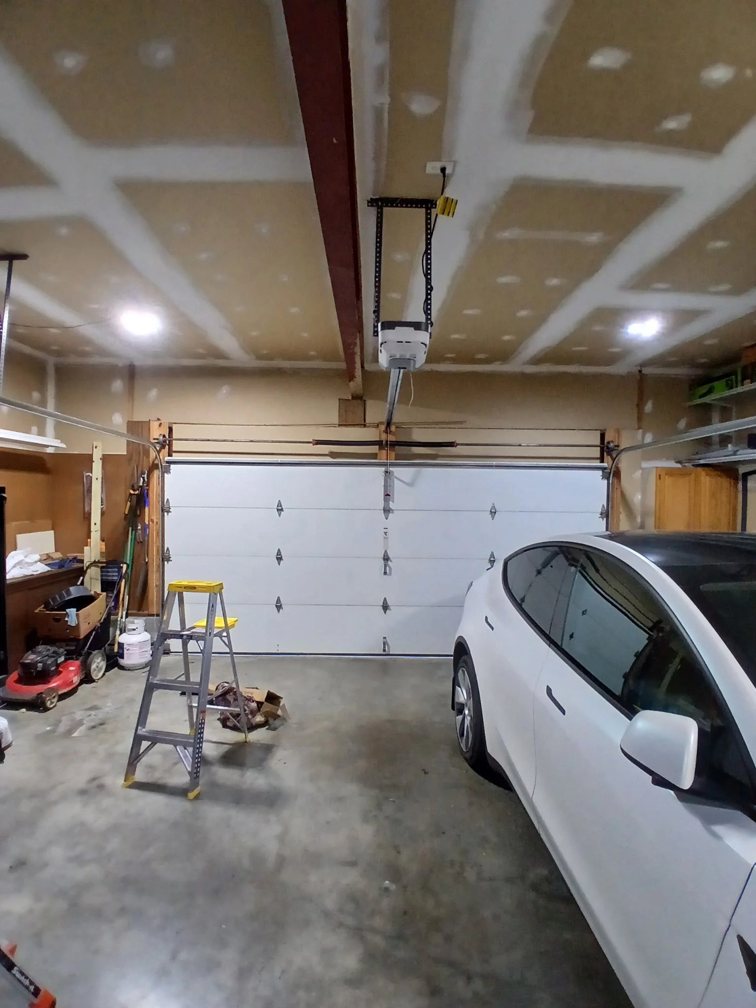View of a garage with a car parked on the right, a white garage door, a stepladder, tools, and storage shelves. The ceiling has drywall with joint compound applied to seams. This was a spring replacement.