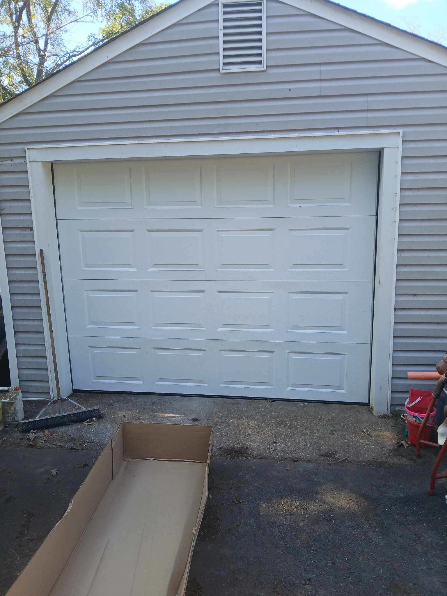 A white garage door on a house with vinyl siding. An empty cardboard box is placed on the ground in front of the garage. To the right, there is a gardening toy and a red bucket. This was a garage door installation.