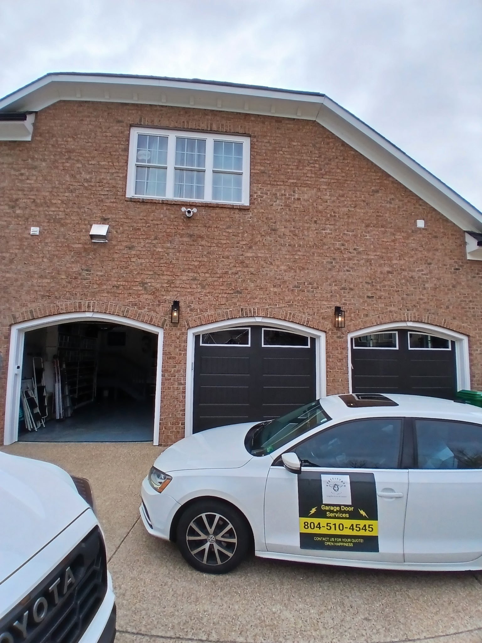 White car with Garage Door Services sign parked in driveway of brick house with black garage doors and a second story window. This was a 3 door garage door opener install job.