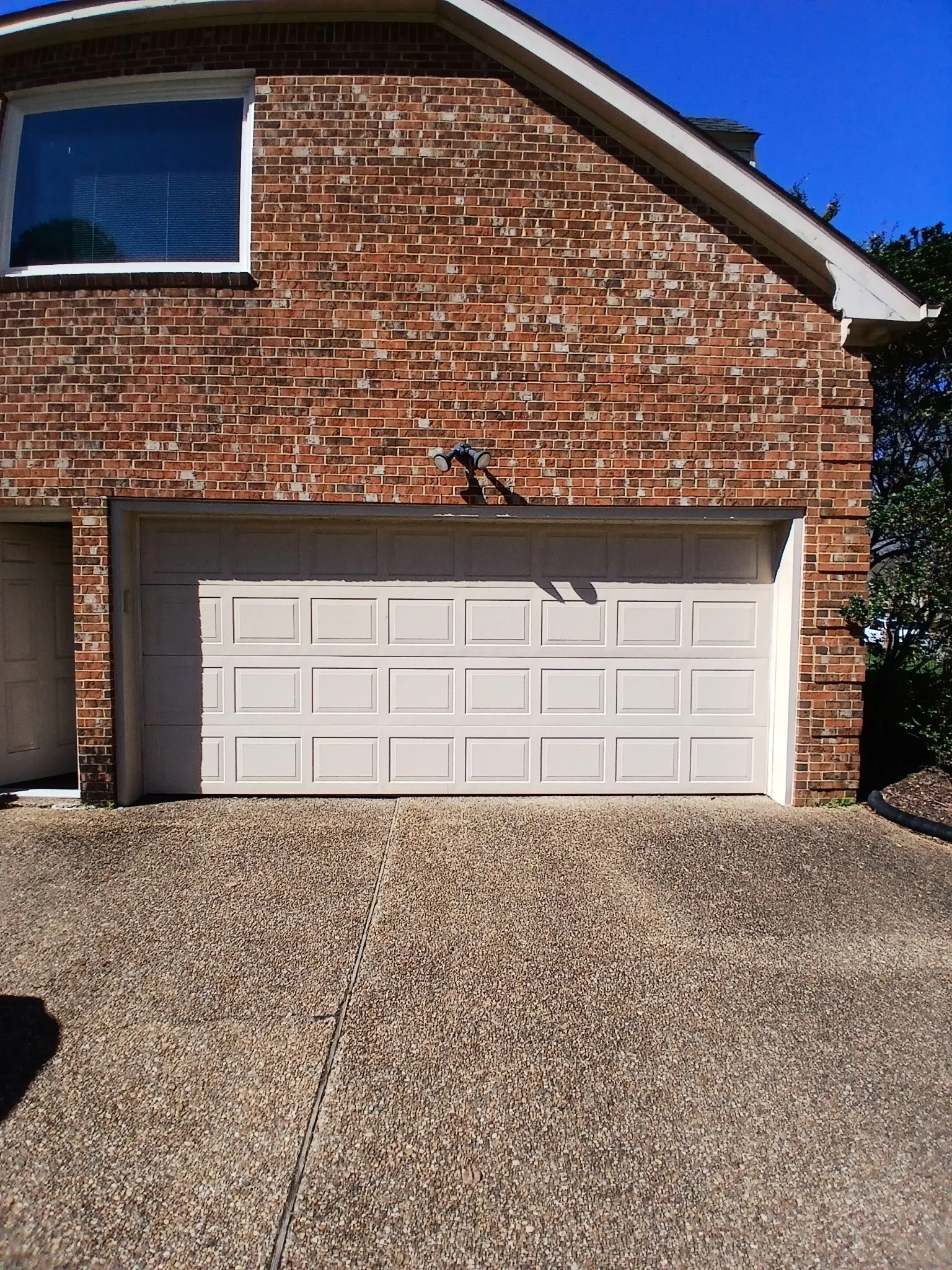 View of a garage with closed white doors, brick exterior, and a window on the upper left. There are outdoor lights above the garage door and trees on the right side. The driveway is textured concrete. This was a garage door installation.