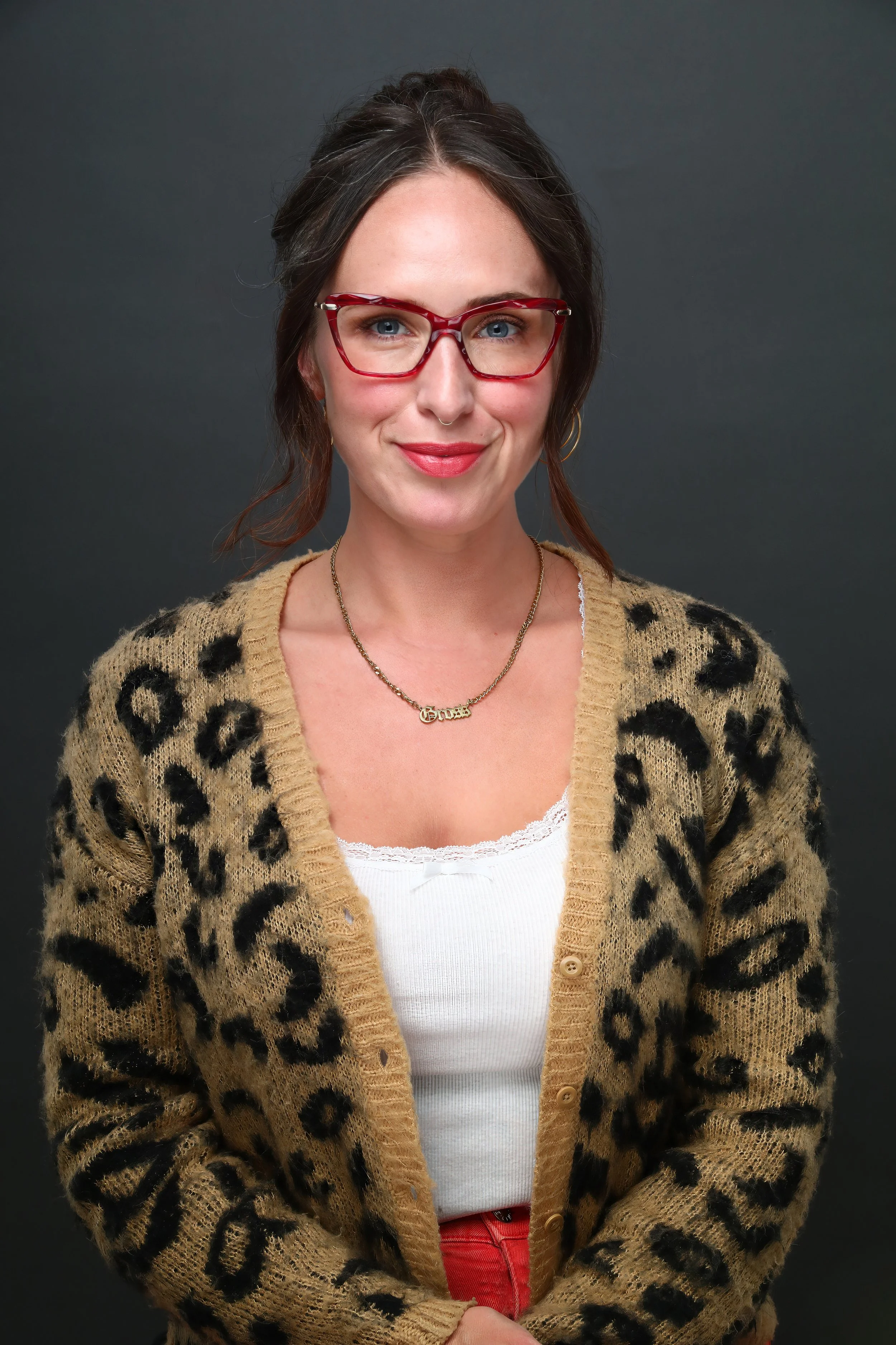 A woman with dark hair, blue eyes, glasses, and a nose ring, wearing a leopard print cardigan over a white top and red pants, standing against a dark background.