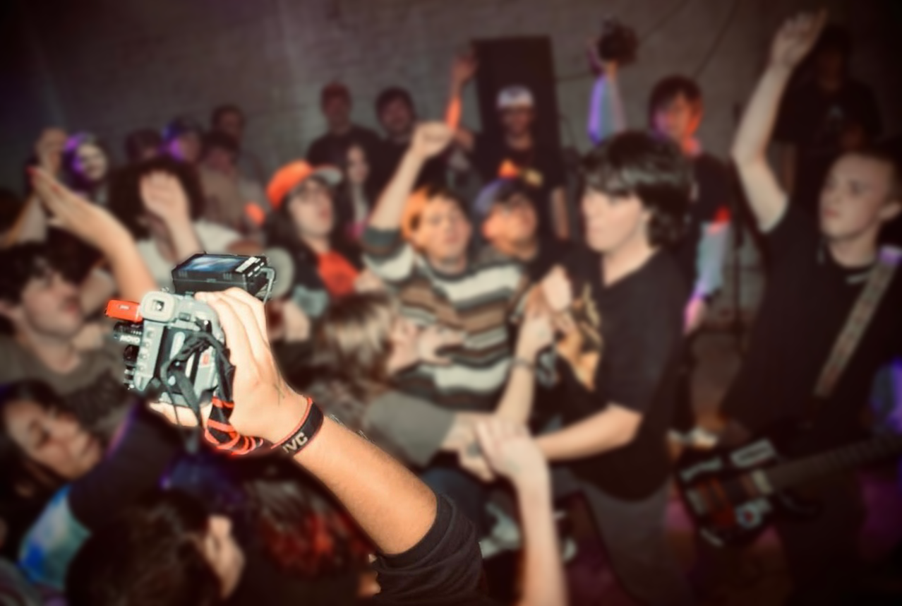 A crowd of people at a concert or party with a woman holding a camera in the foreground.
