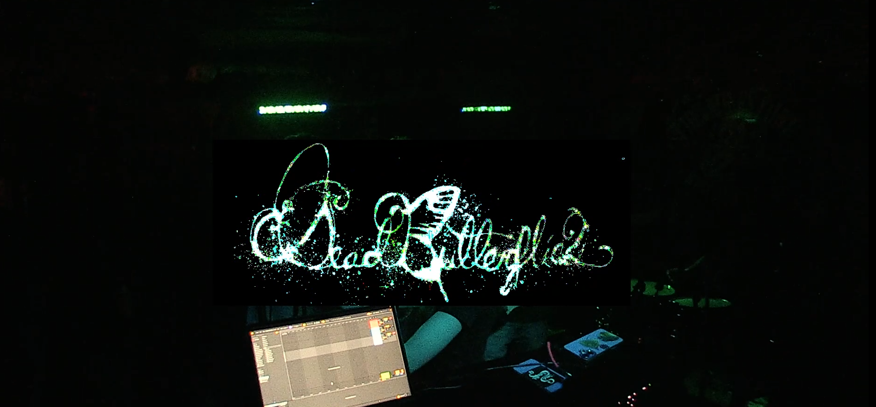 Dark room with a digital screen displaying the words 'Dad and Grandpa' in stylized, illuminated script, surrounded by splatter effects. A laptop with a music production software is visible in front of the screen, and some electronic devices and a phone are also present.