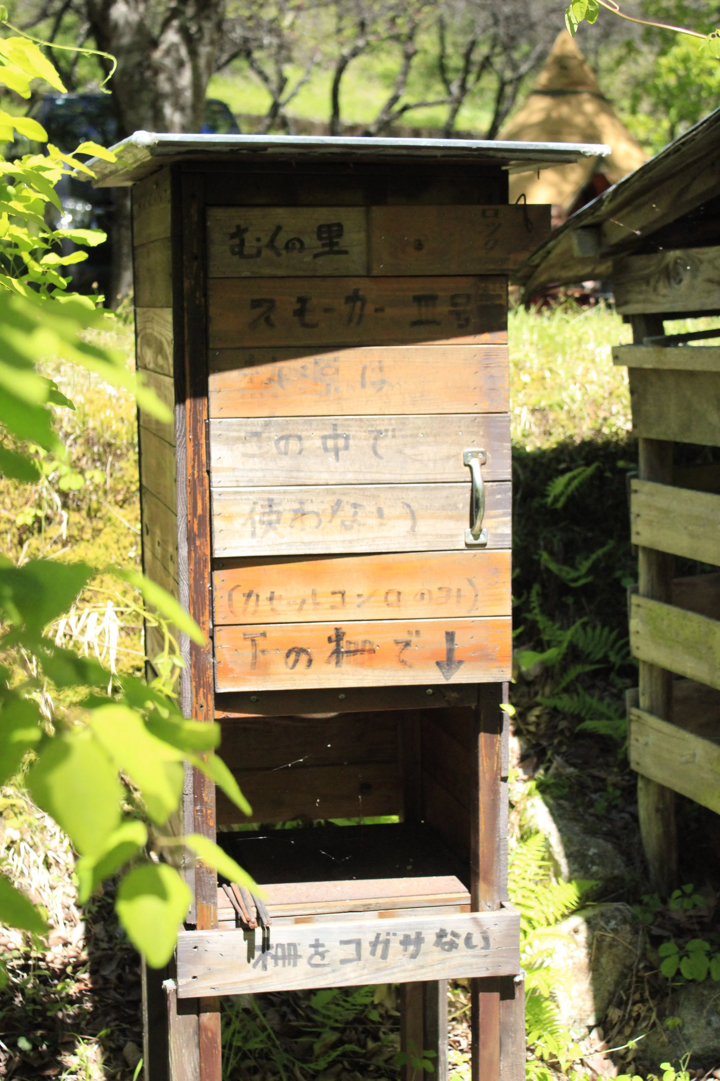 木製の看板と木製の箱が屋外の自然の中に設置されている。看板には日本語で書かれた文字が見えるが、読むのが難しい。背景には木や草、葉などの植物が見える。