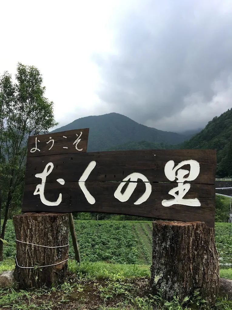 山と雲の背景に木製の看板があり、白い文字で「ようこそむくの里」と書かれている。