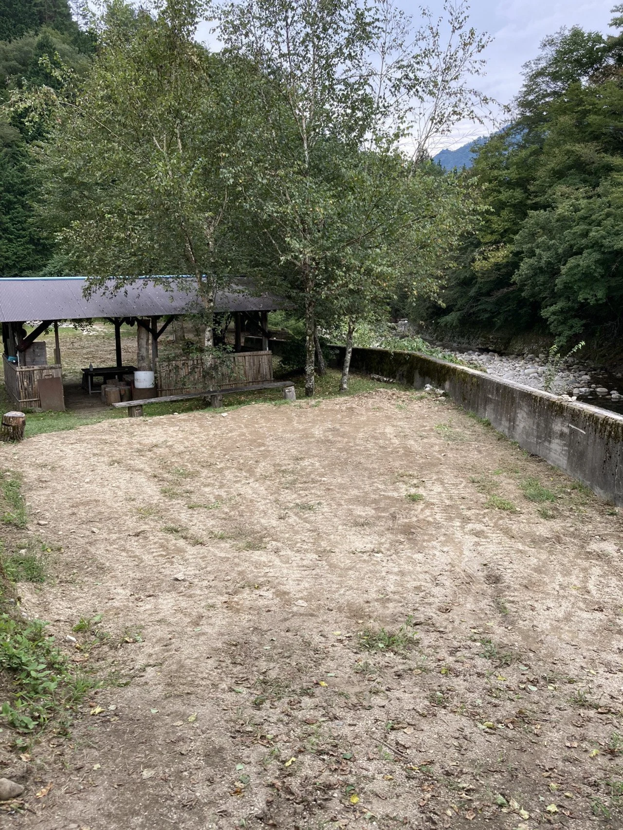 自然の中の広い土の空き地、左側に木製の休憩所、背景には木と山と空が見える風景