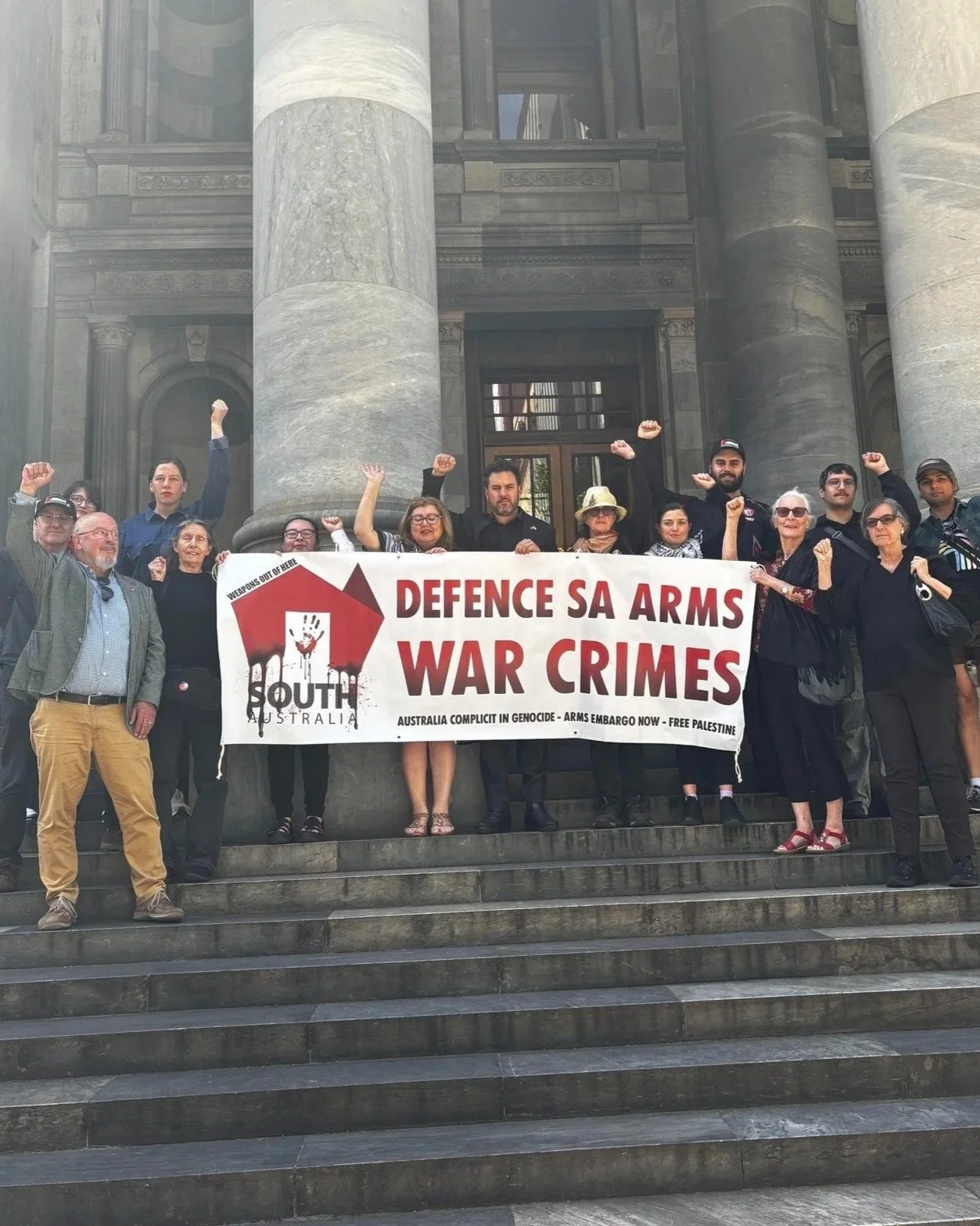 A group of diverse people standing on the steps of SA Parliament, holding a protest banner that reads "DEFENCE SA ARMS WAR CRIMES" . They are raising clenched fists in solidarity.