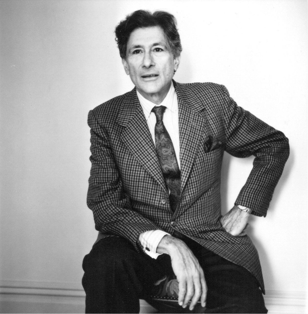 Black and white photograph of Edward Said sitting on a chair, wearing a checkered blazer, white shirt, and patterned tie, with one arm resting on his knee and the other hand on his hip.