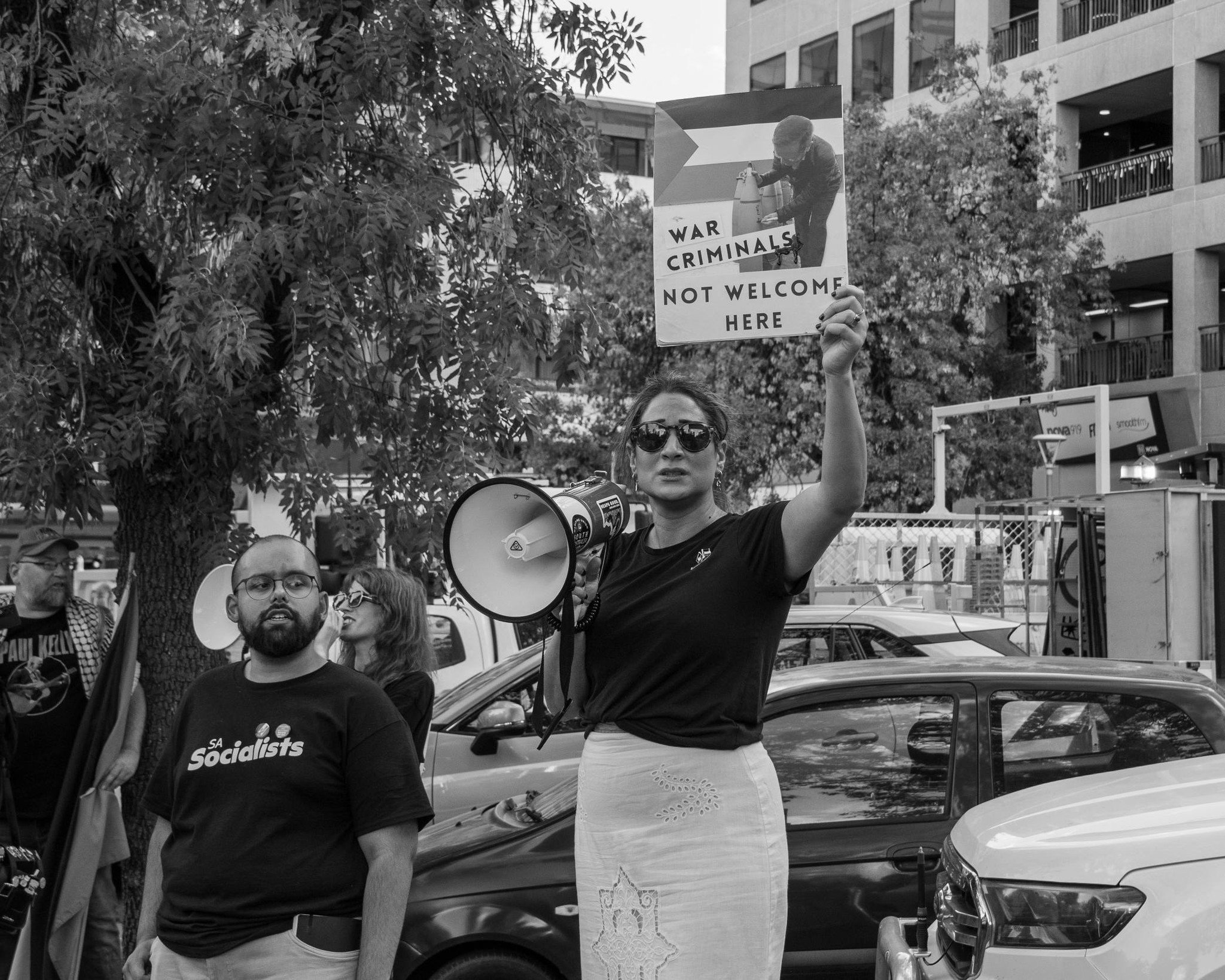 A woman wearing sunglasses holding a sign at a protest in Adelaide. The sign shows a graphic of a person attaching bombs to a rocket launcher with the words "WAR CRIMINALS NOT WELCOME HERE."