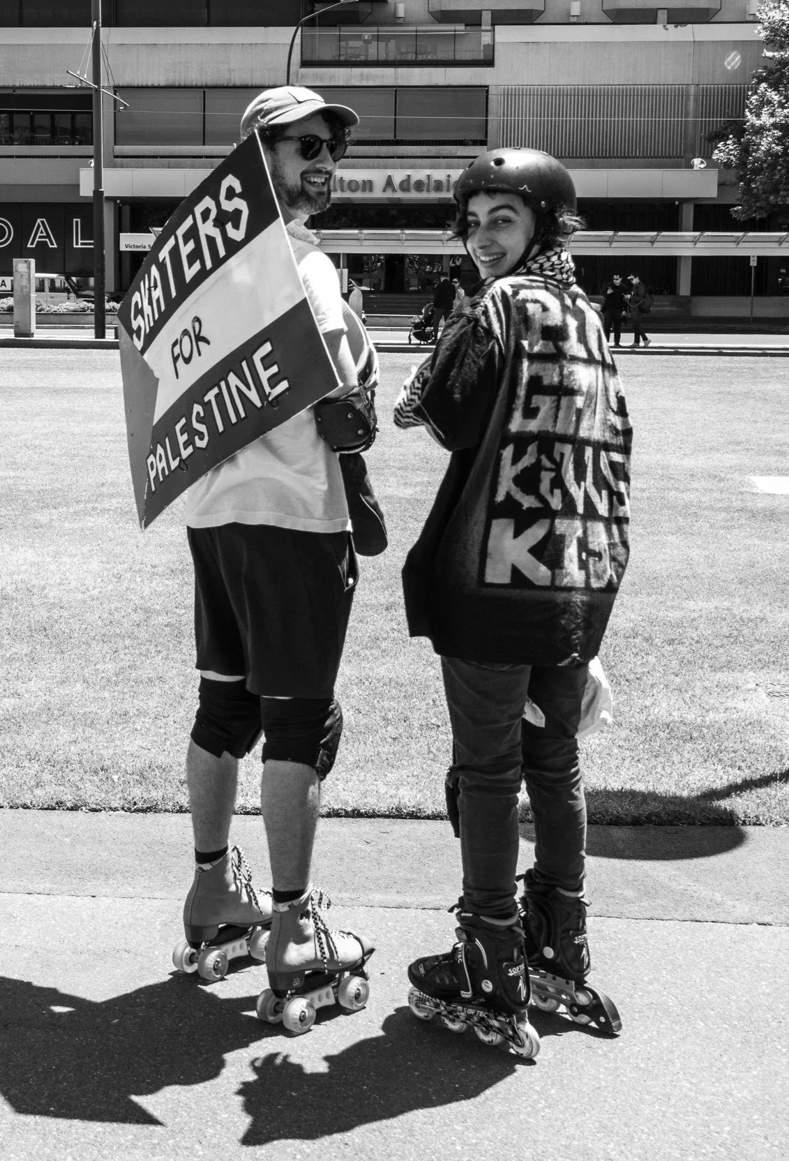 Two people in roller skates and helmets smiling at each other on a sidewalk in front of a building. One person is holding a sign that reads 'Skaters for Palestine.'  Image taken by Vicky Dennison.