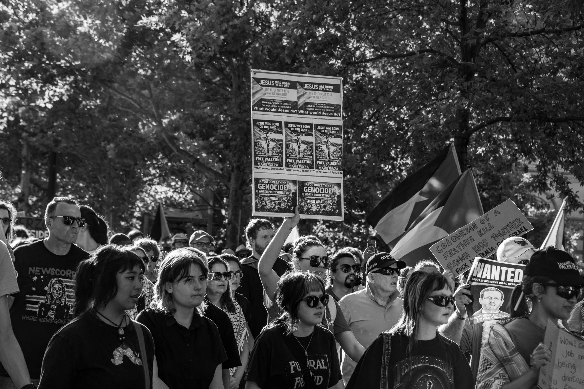 A group of people gathered in a protest or rally outdoors with trees in the background. Individuals are holding signs and banners. Image taken by Vicky Dennison.