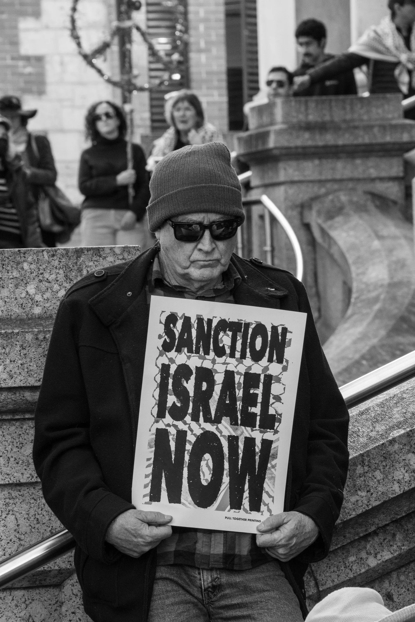 A man in dark clothing, a beanie, and sunglasses is sitting on a bench, holding a protest sign that reads 'Sanction Israel Now'. He has a serious expression. In the background, several people are standing.  Image taken by Vicky Dennison.