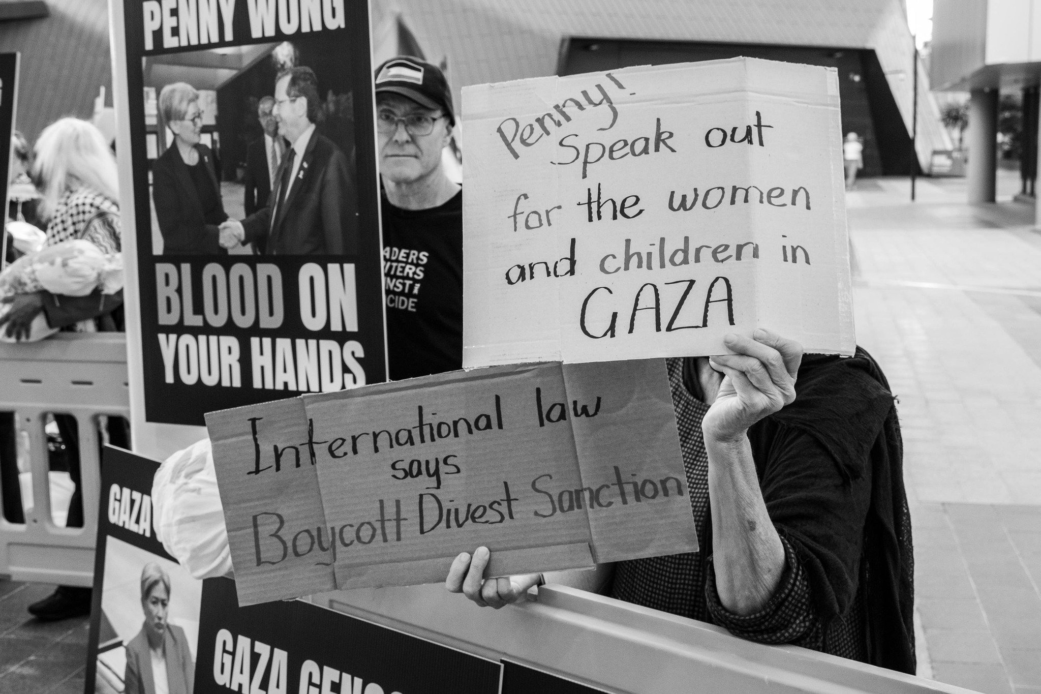 A person holding a cardboard sign with handwritten text that says 'Penny! Speak out for the women and children in Gaza'. In the background, there are posters and other people at a protest or rally. Image taken by Vicky Dennison.