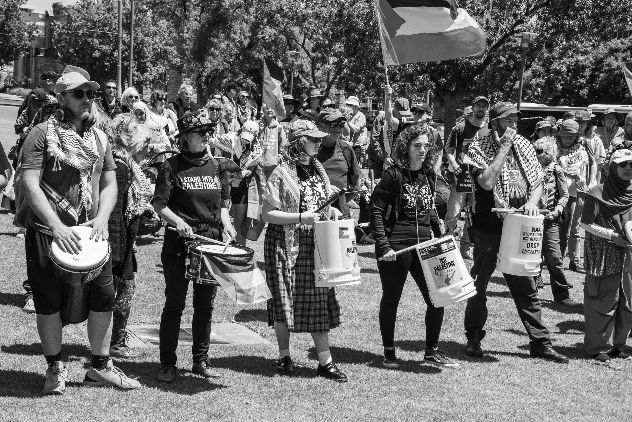 Black and white photo of a diverse group of people participating in a protest or rally. Many hold signs and flags, some wear shirts with messages advocating for Palestine.  Image taken by Vicky Dennison.