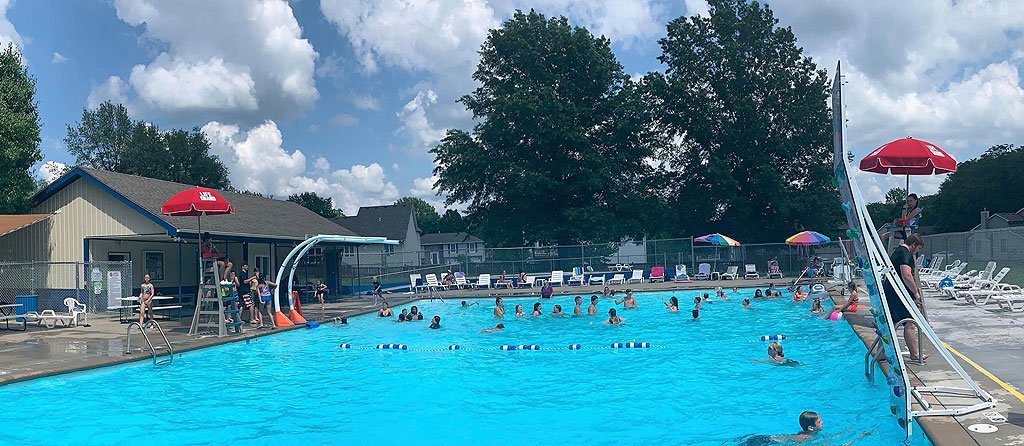 Public outdoor swimming pool with multiple people swimming and playing, surrounded by colorful umbrellas and lounge chairs, with a small building and a tree in the background.