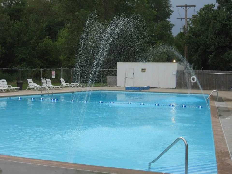 Empty outdoor swimming pool with water spray and lounge chairs, surrounded by trees and fencing.