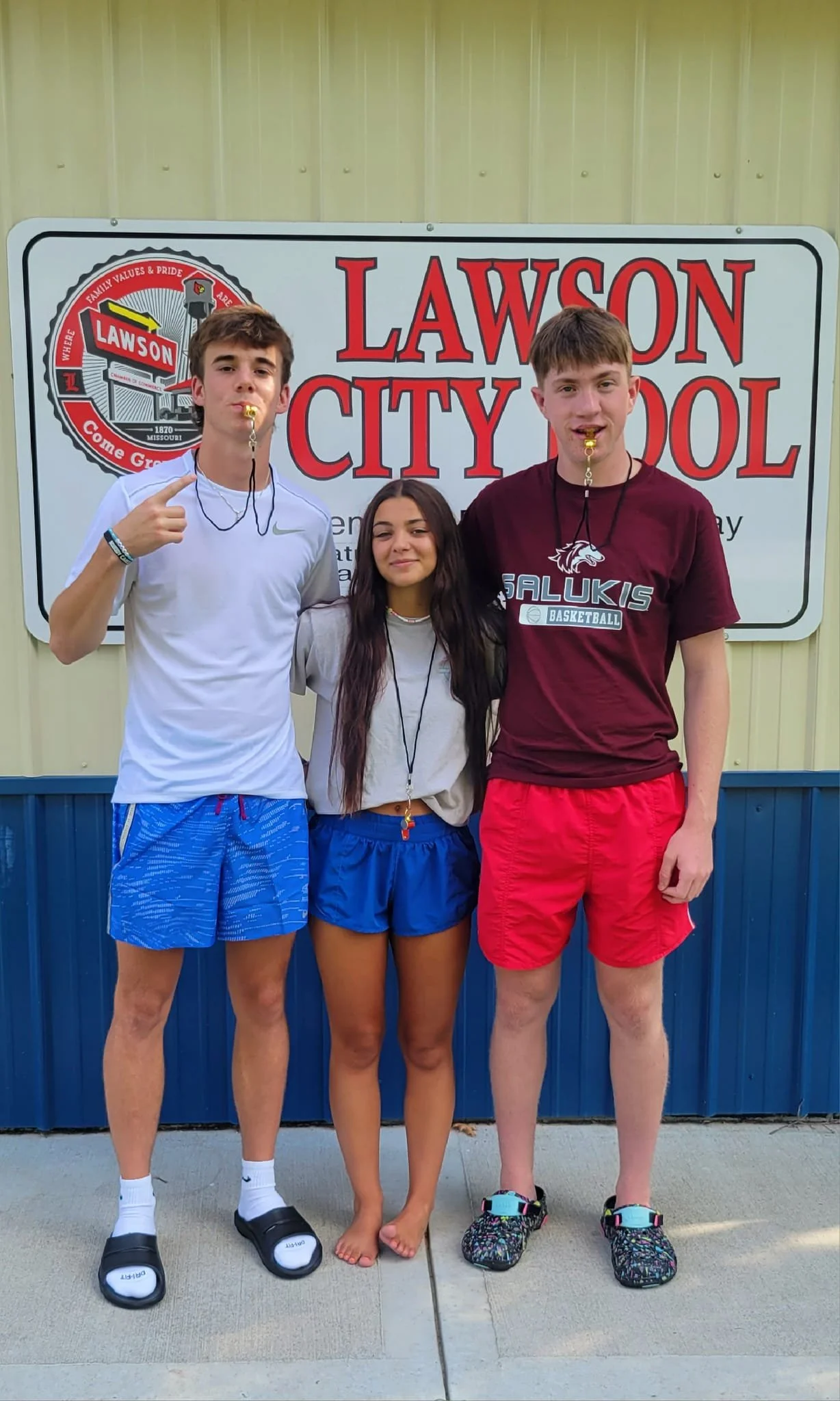 Three teenagers standing in front of a sign that reads "Lawson City School." The boy on the left is wearing a white t-shirt, blue shorts, and slides. The girl in the middle is wearing a beige t-shirt and blue shorts, barefoot. The boy on the right is wearing a maroon t-shirt with "Salukis Basketball" and red shorts, holding a whistle in his mouth.