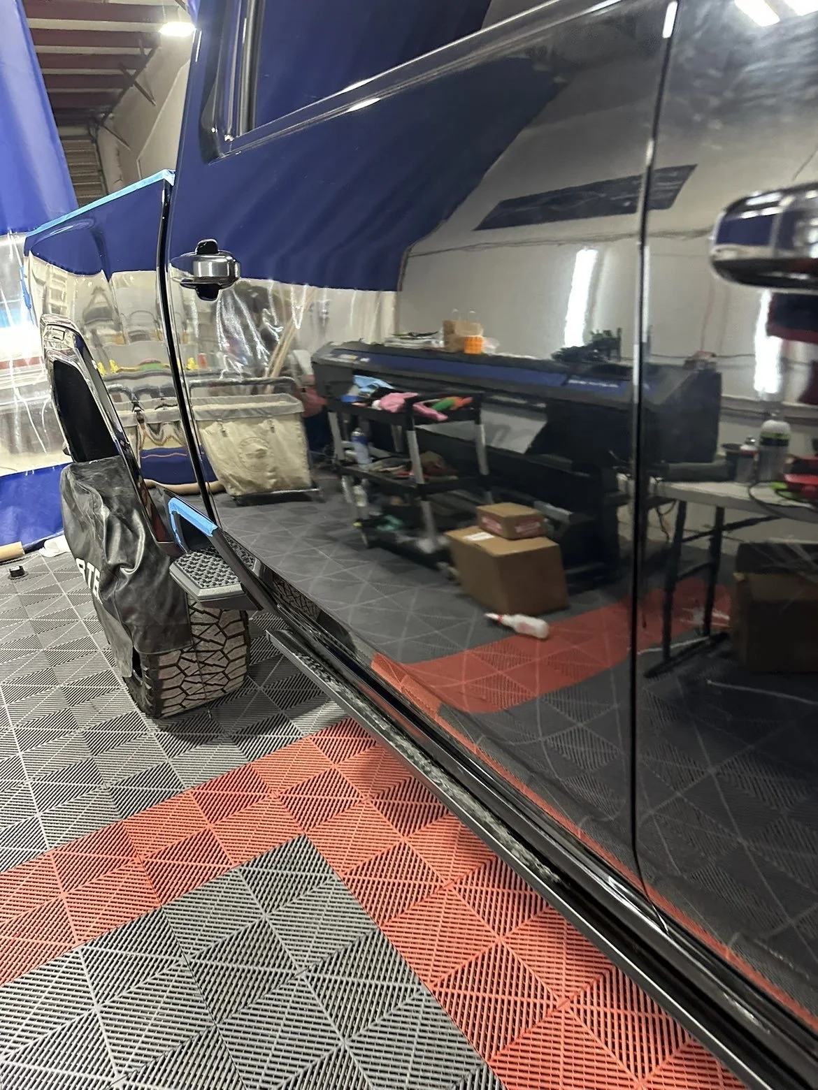The photo shows a garage or workshop with a black car's side reflecting the room. Reflected in the car are tools, boxes, a keyboard, and other workshop items. The floor has a black and orange checkered mat.