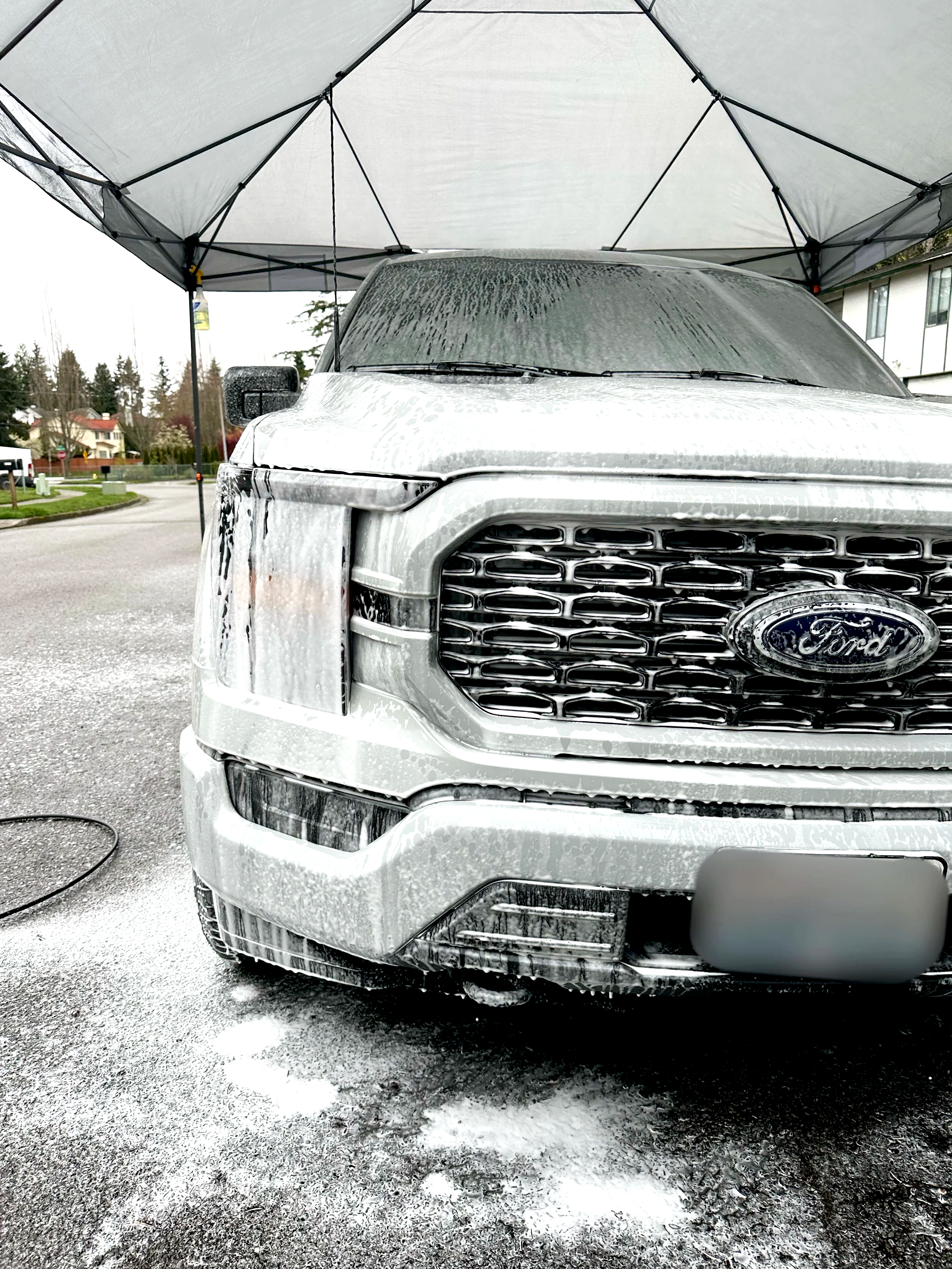 Front of a Ford vehicle being washed with foam under a gray canopy, in a residential neighborhood.