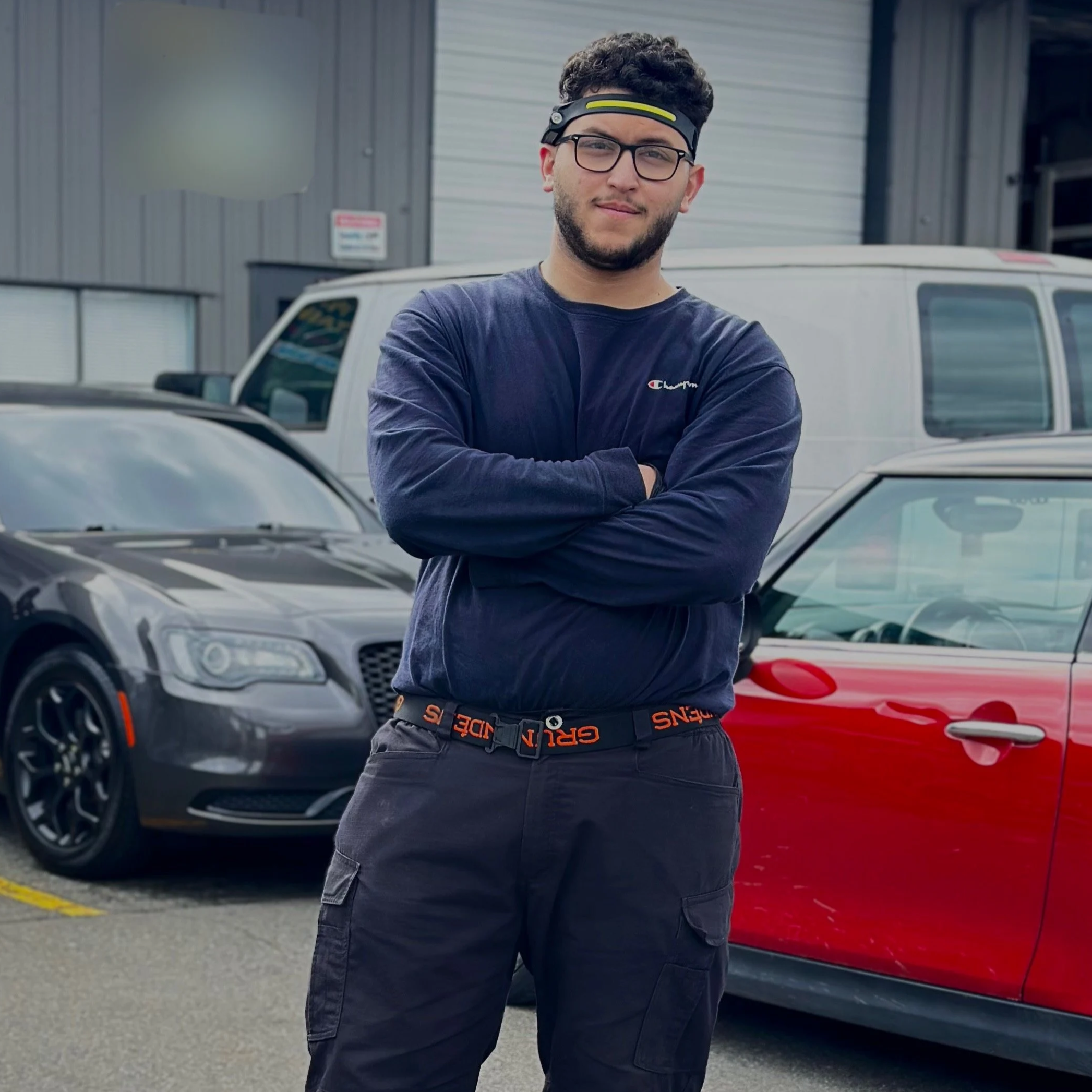 A young man with glasses and curly hair standing with arms crossed in a parking lot, wearing a headband, a dark long-sleeve shirt, and cargo pants, with several parked cars, including a black car and a red vehicle, and a gray building in the background.
